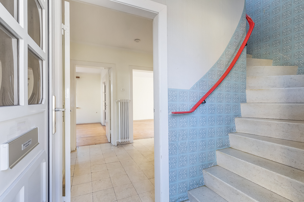 Eingangsbereich mit Fliesenboden, offener Tür, Treppe mit rotem Geländer und Radiator.