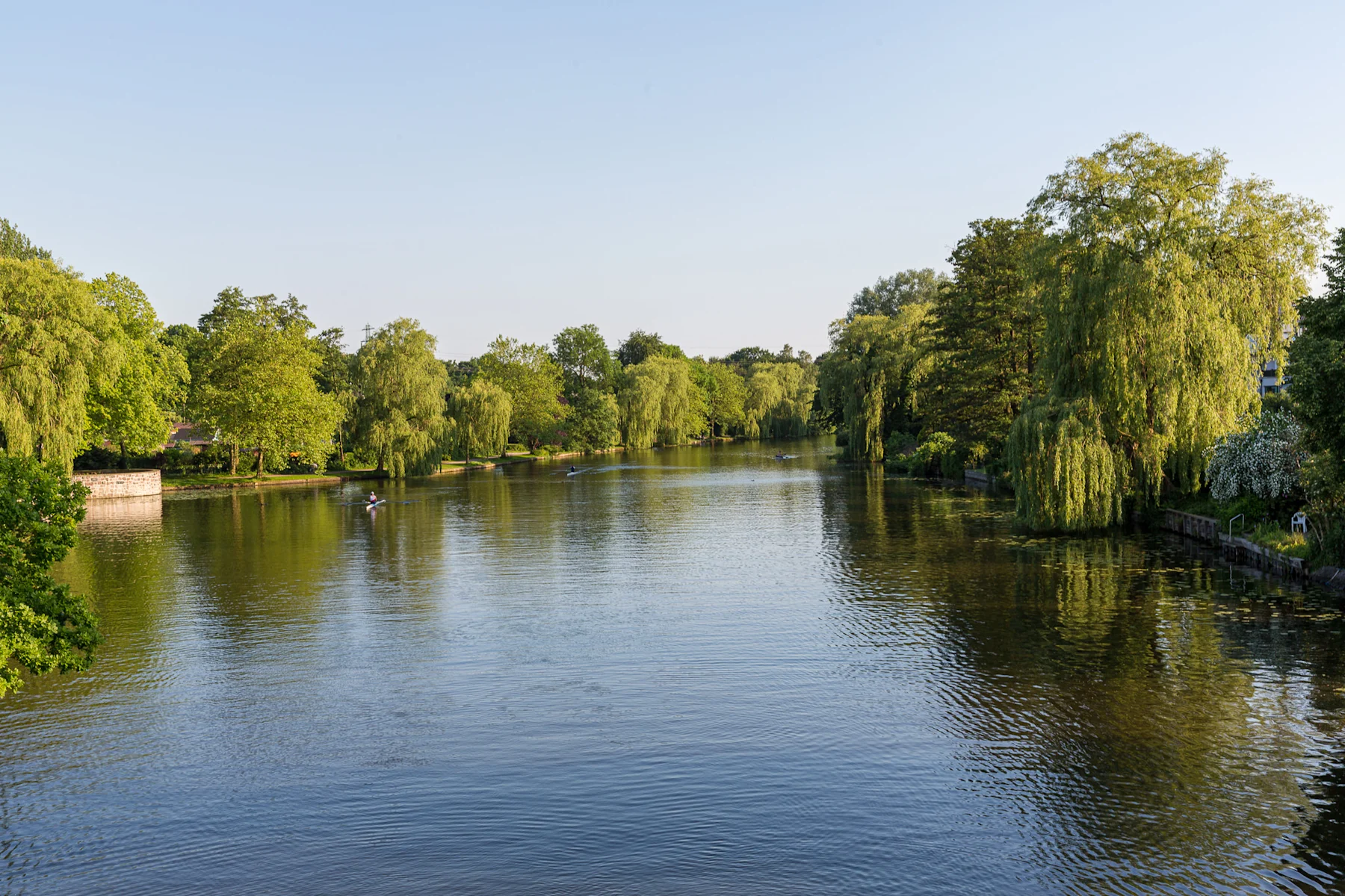 Fluss mit grünem Ufer und Bäumen bei klarem Himmel, einige kleine Boote auf dem Wasser.