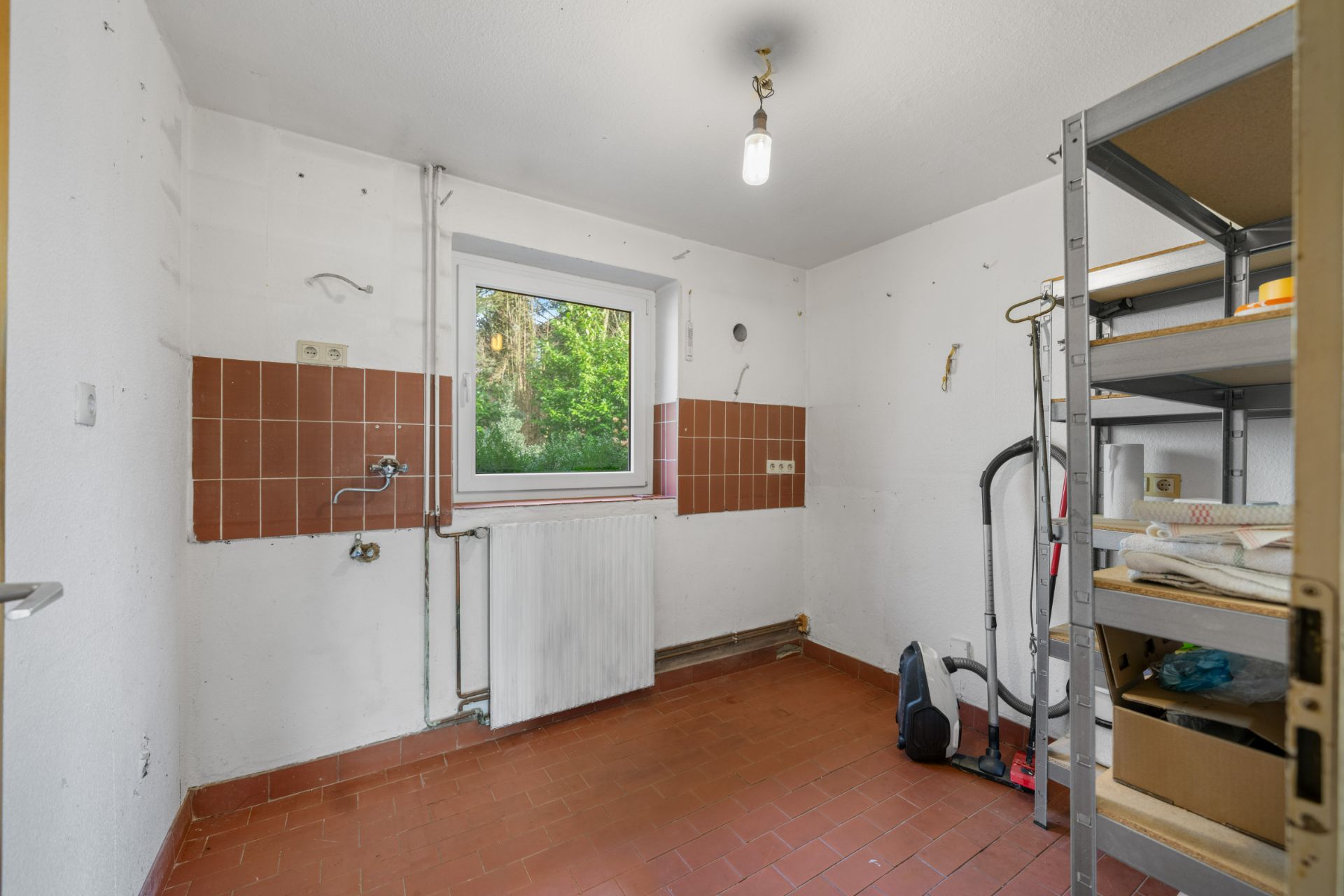 Leerer Raum mit braunen Fliesen, Regal, Fenster und Reinigungsgeräten an der Wand.
