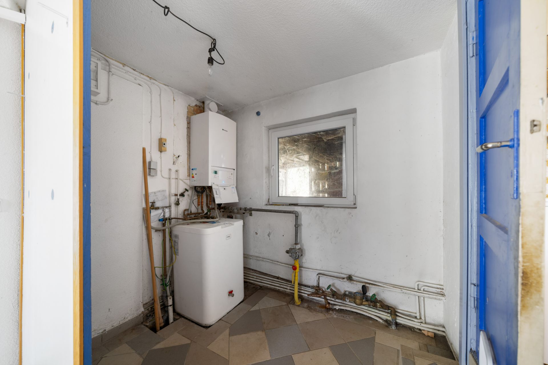 Kleiner Hauswirtschaftsraum mit Heizung, Boiler, Fenster und einigen Rohren an der Wand.