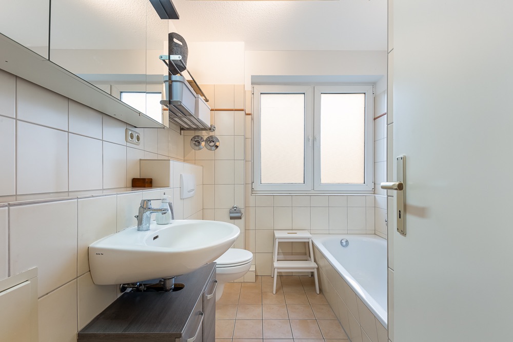 Helles Badezimmer mit Waschbecken, Toilette, Badewanne und zwei Fenstern.