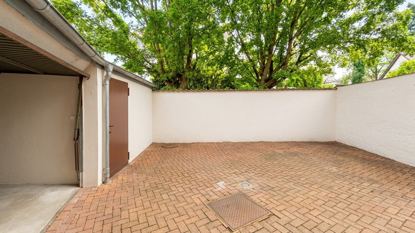 Gepflasterter Innenhof mit Garage und weißen Mauern, im Hintergrund grüne Bäume.
