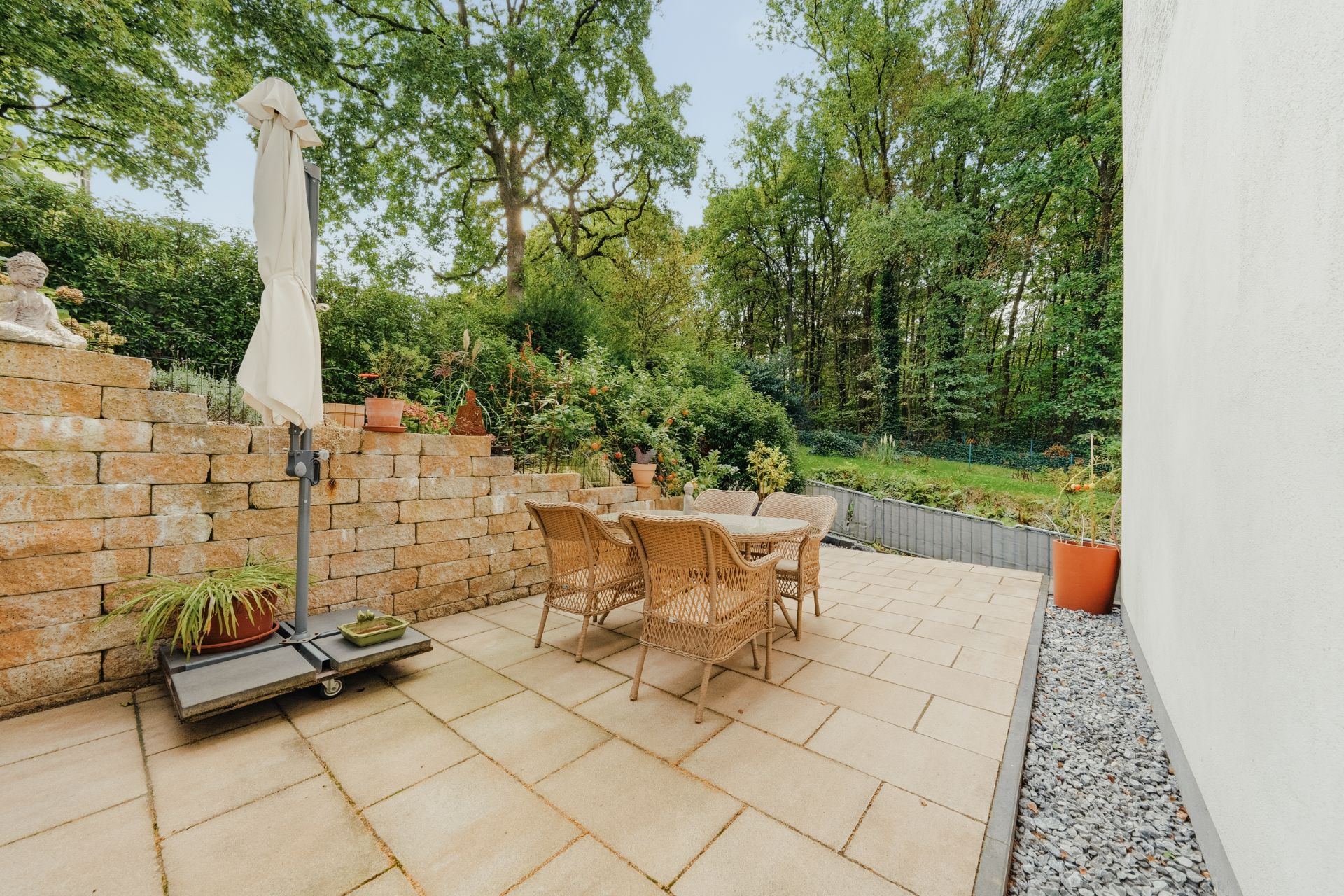 Gepflasterte Terrasse mit Tisch, Stühlen, Sonnenschirm und Pflanzen, neben einer Mauer und viel Grün.