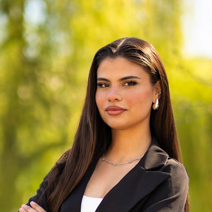 Junge Frau mit langen braunen Haaren und schwarzem Blazer steht draußen und verschränkt die Arme.