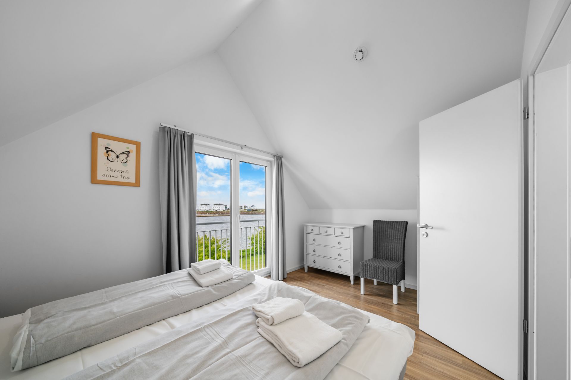 Helles Schlafzimmer mit Doppelbett, Kommode, Stuhl und Fenstern mit Blick nach draußen.