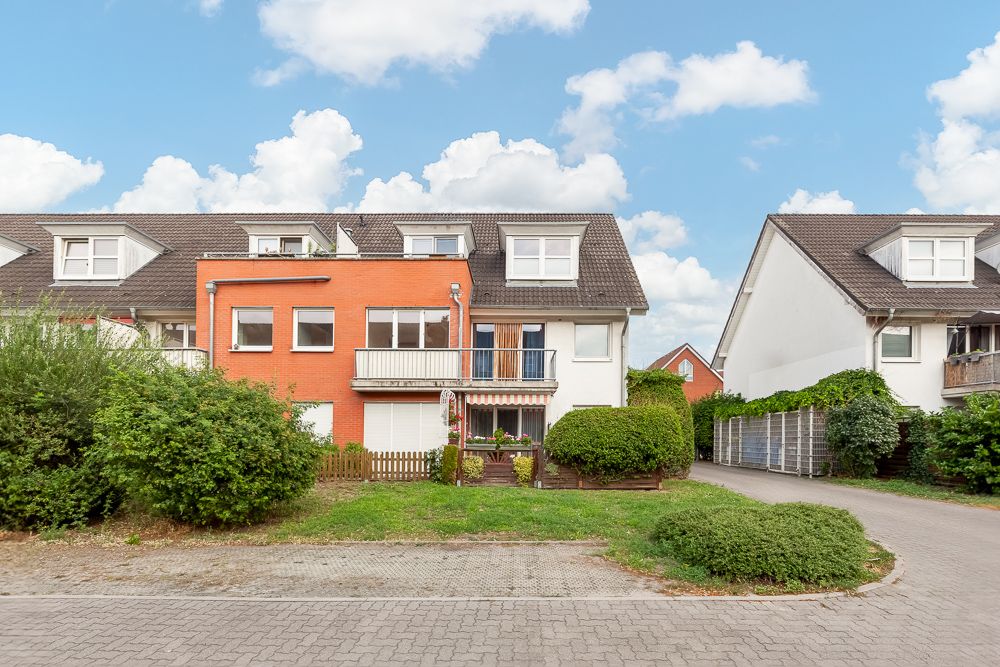 Modernes Wohnhaus mit rotem Anbau, Balkon, Garten und gepflegten Büschen vor dem Gebäude.