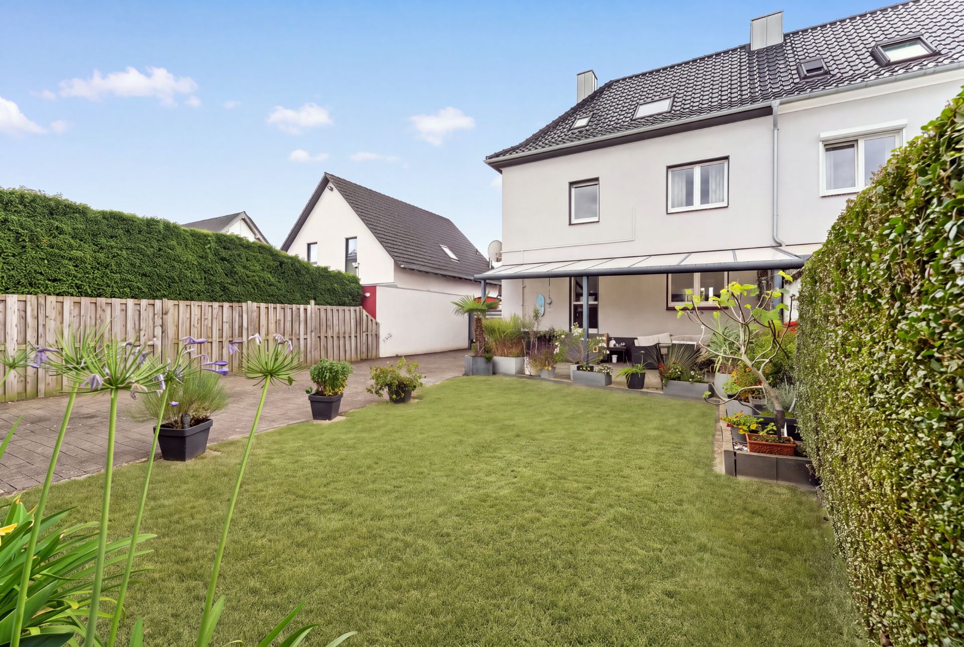 Grüner Garten mit Terrasse an einem weißen Wohnhaus, viele Pflanzen und Rasenfläche.