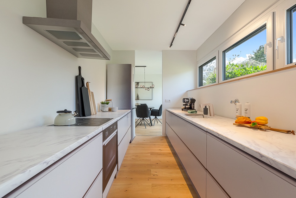 Moderne Küchenzeile mit weißen Arbeitsplatten und großen Fenstern, Blick ins Esszimmer.