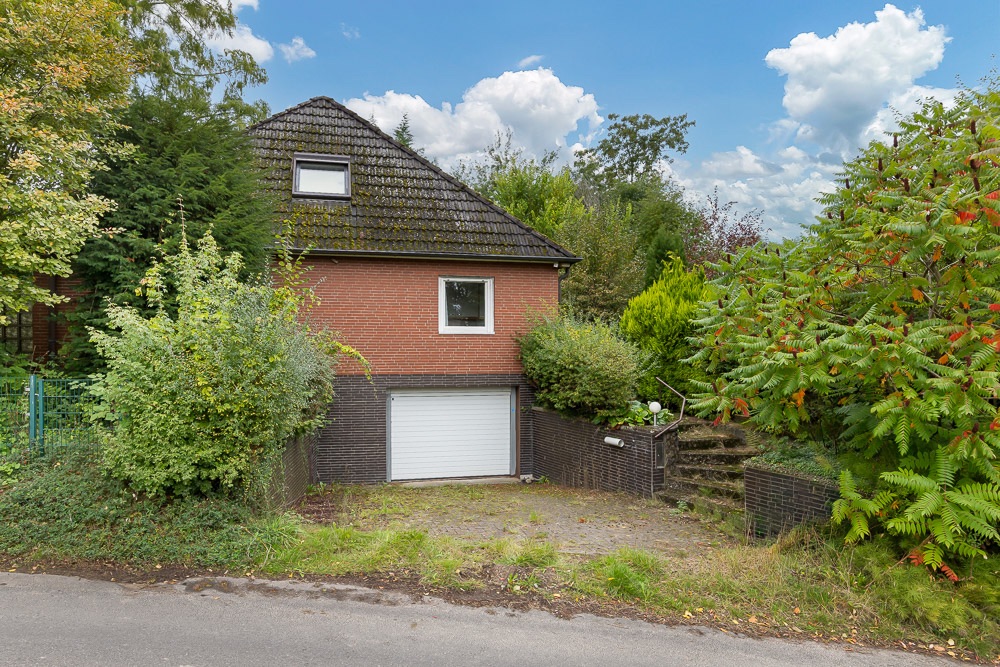 Rotes Einfamilienhaus mit Garage, schrägem Dach und umgeben von viel Grün und Sträuchern.