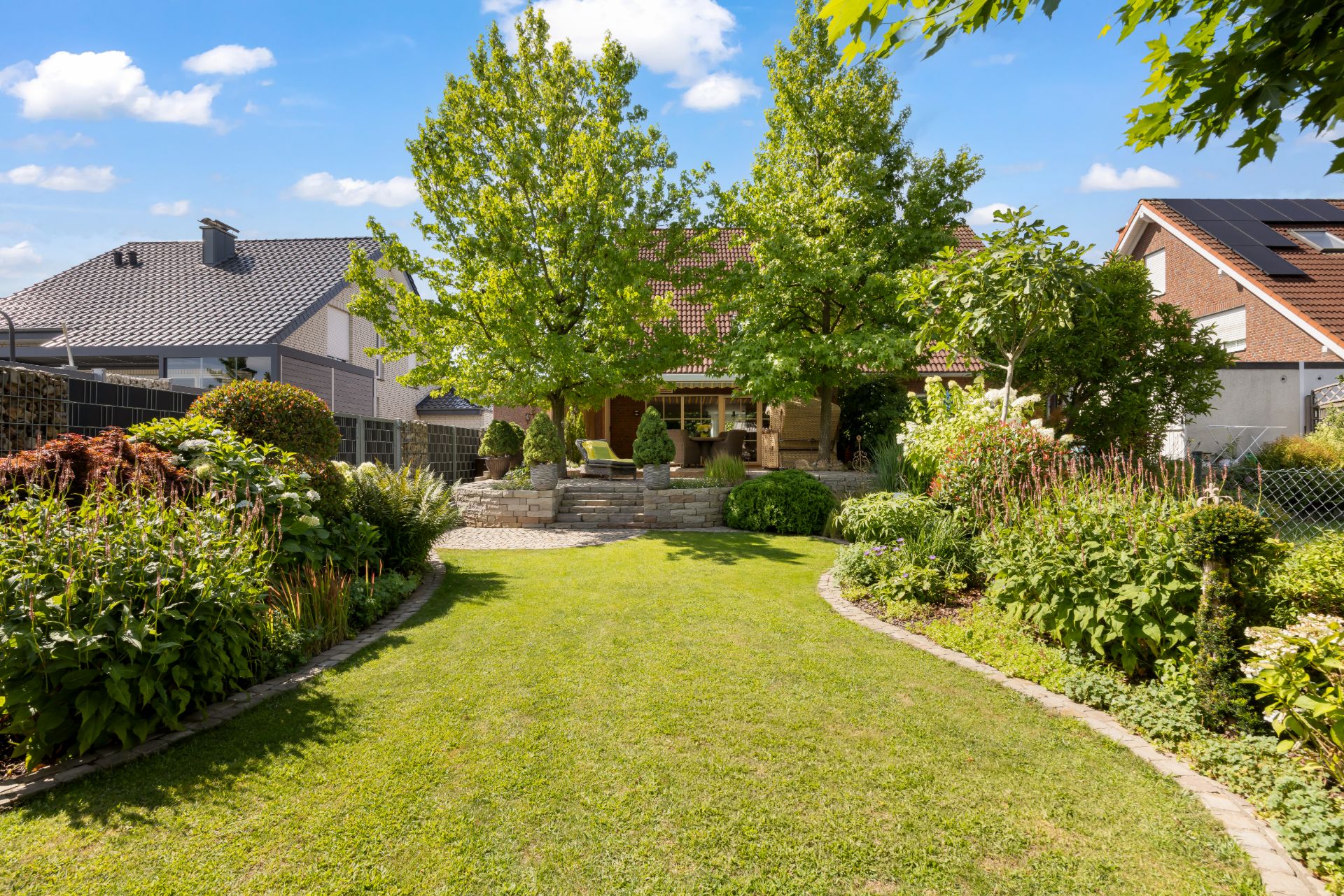 Gepflegter Garten mit Rasenfläche, Bäumen und Blumenbeeten, Haus im Hintergrund.