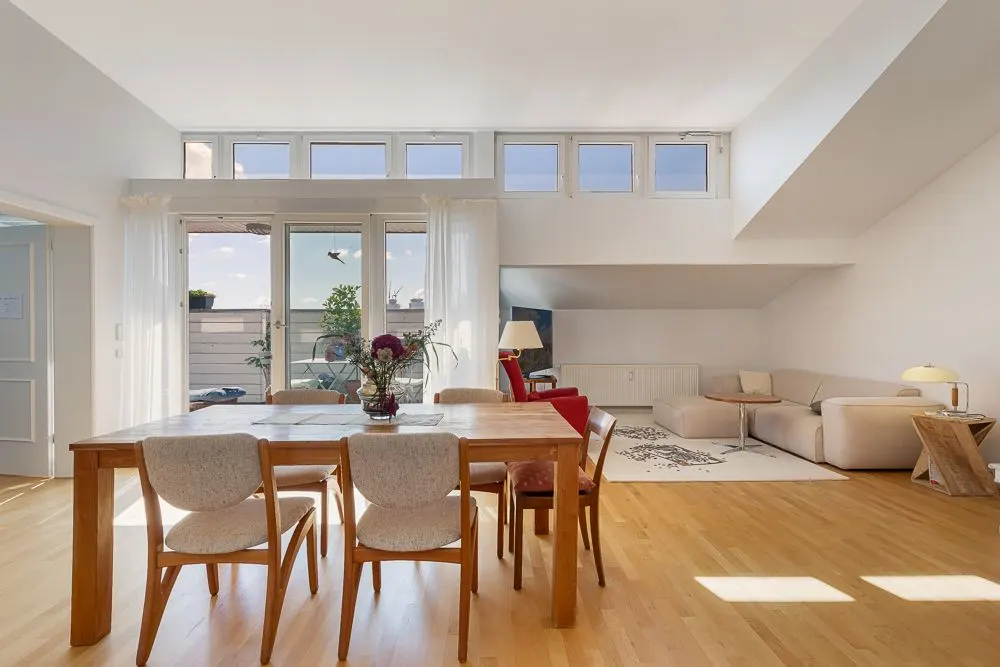 Helles Wohnzimmer mit Holzboden, Esstisch, Stühlen, Sofa und großen Fenstern.