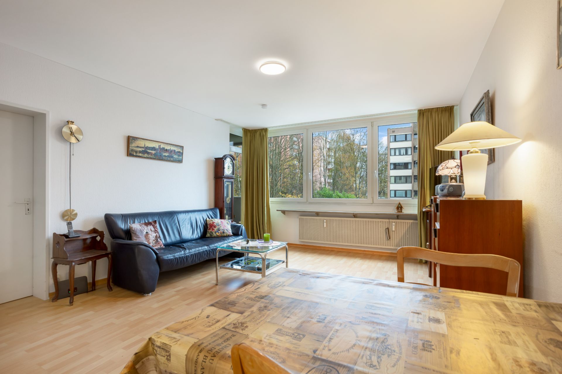 Helles Wohnzimmer mit schwarzem Sofa, Esstisch und großem Fenster mit Blick nach draußen.