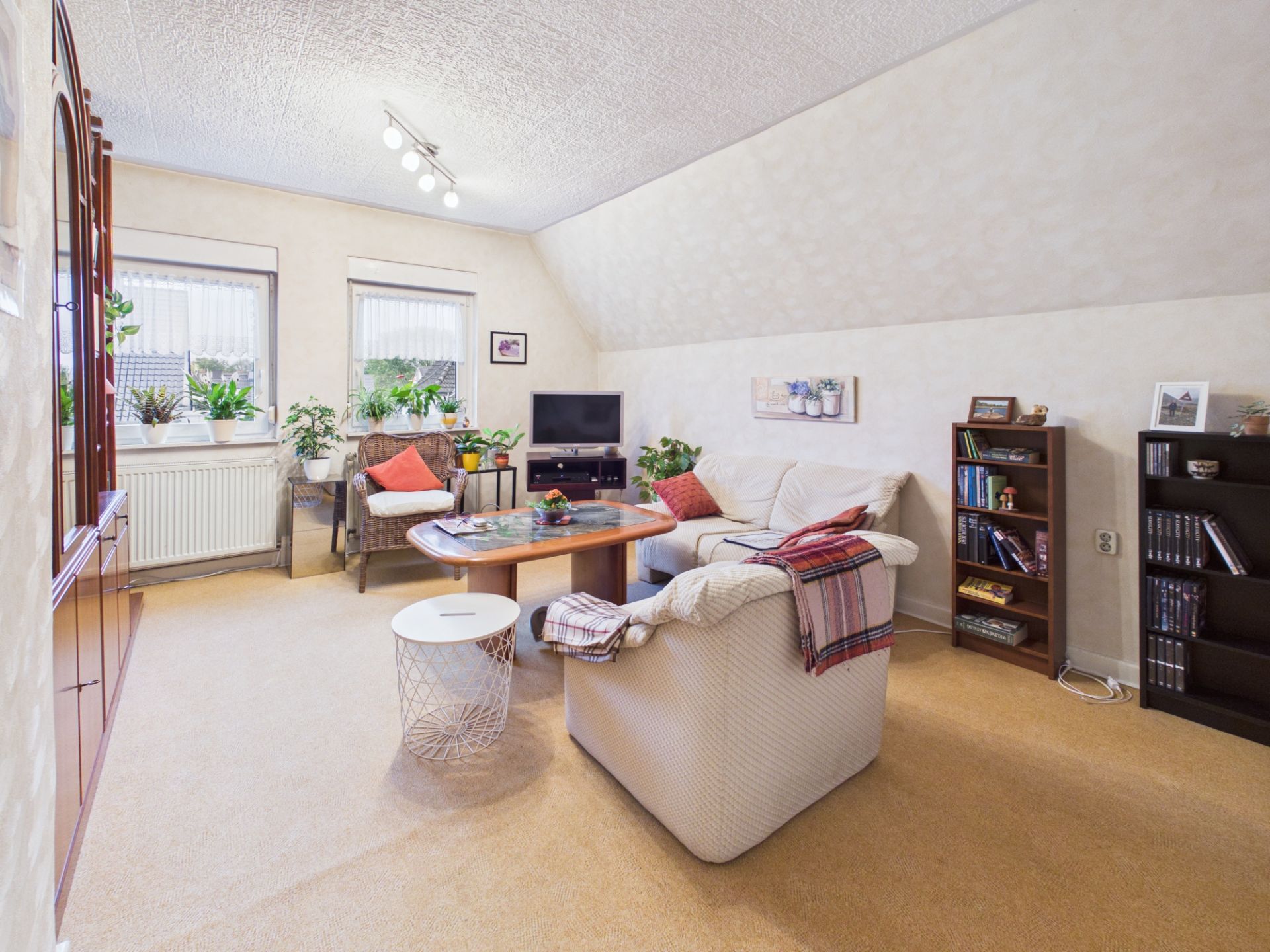 Helles Wohnzimmer mit Sofa, Sesseln, Couchtisch, Bücherregalen und vielen Pflanzen am Fenster.