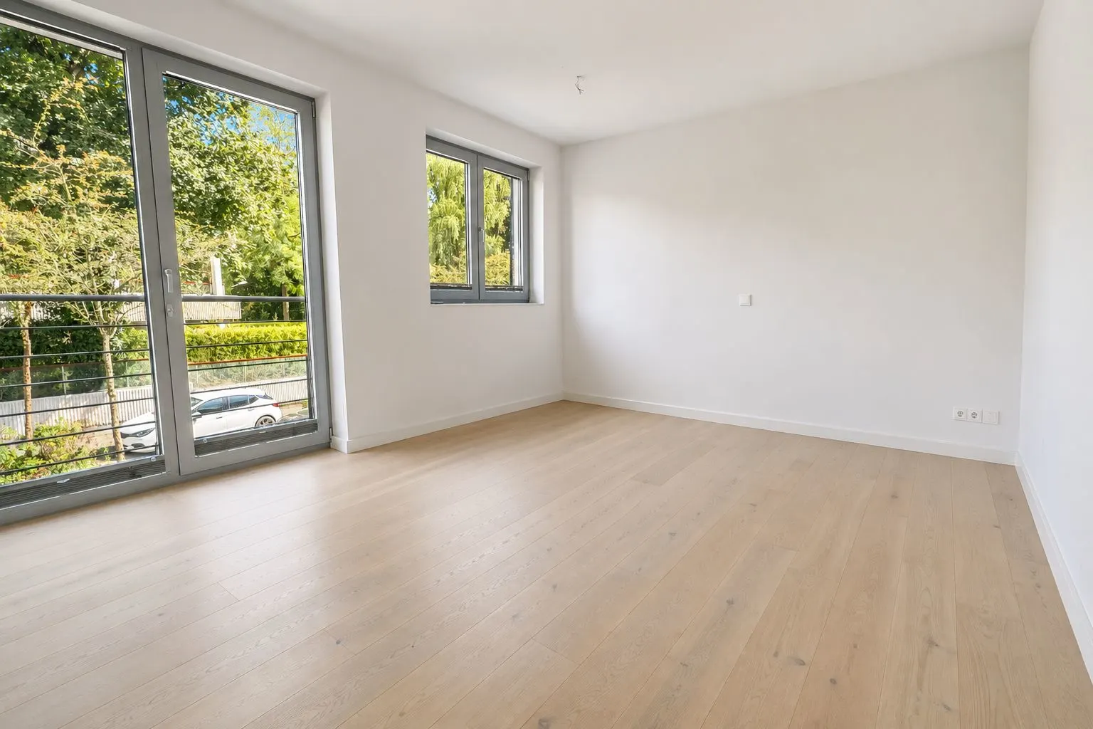 Helles, leeres Zimmer mit großen Fenstern und hellem Holzboden.