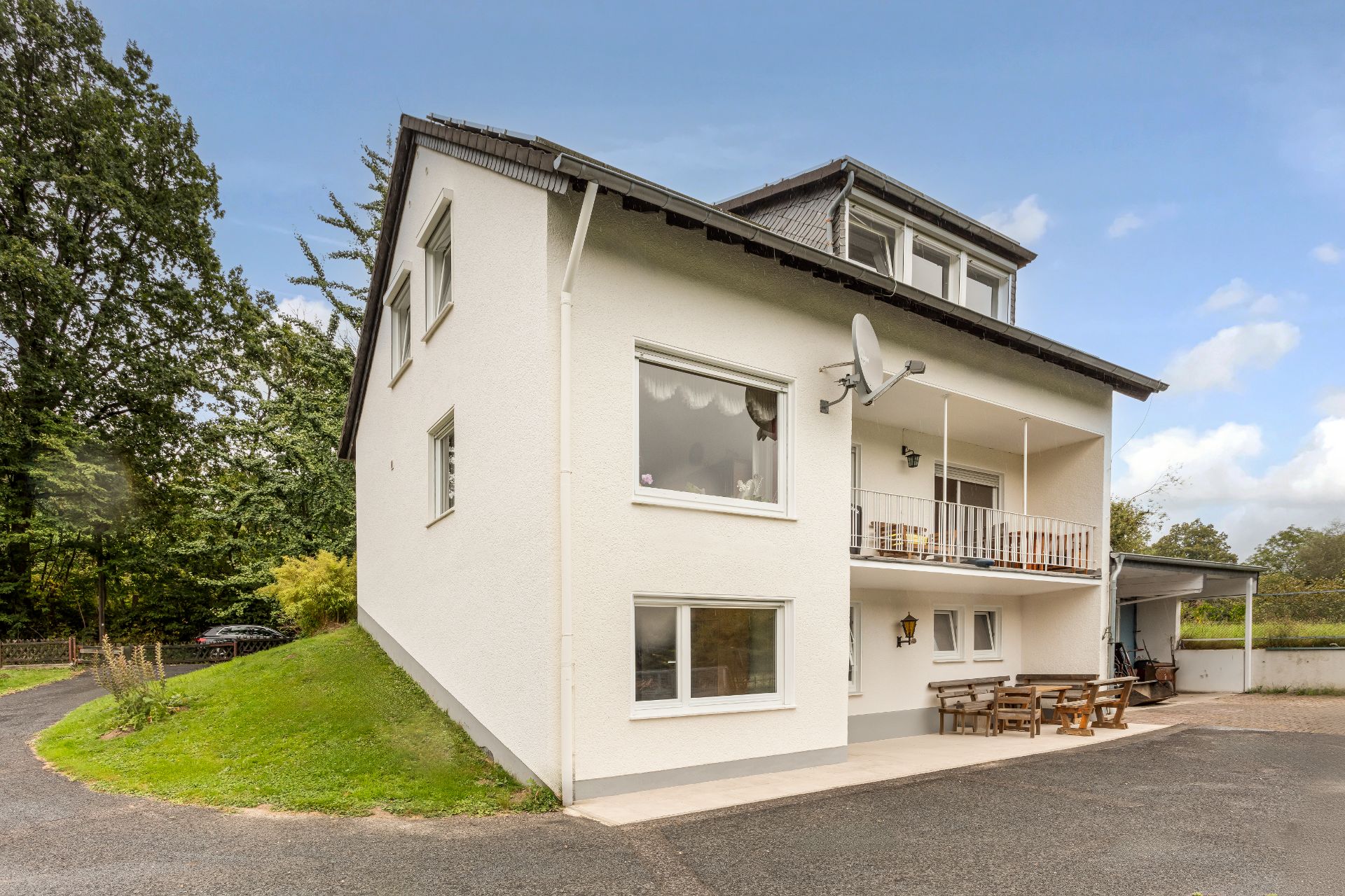 Weißes Wohnhaus mit Balkon, Holzbank und Tischen im Außenbereich, umgeben von Bäumen.