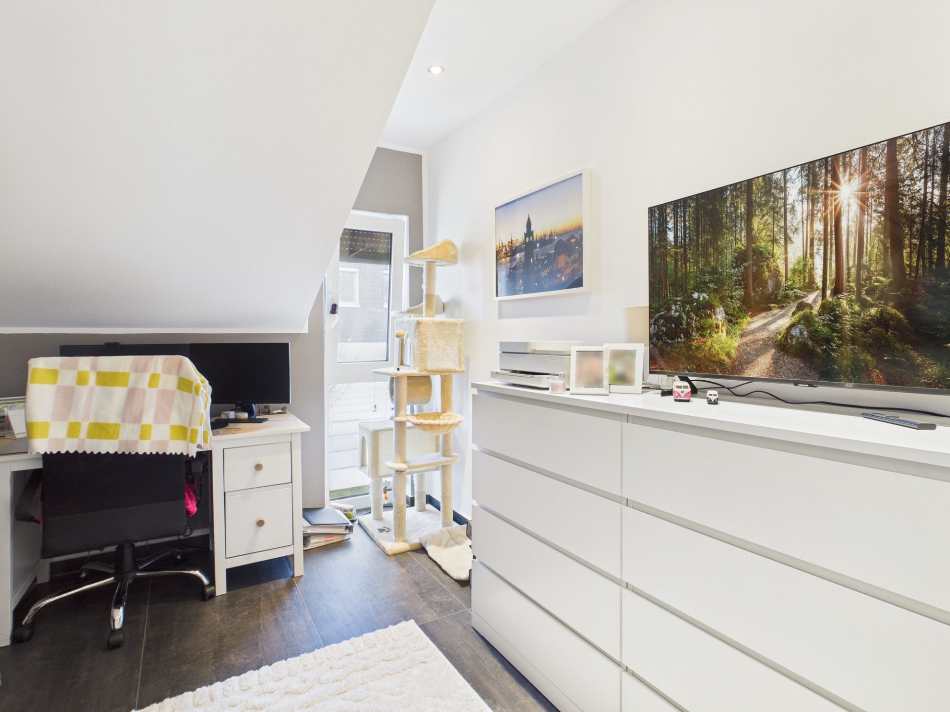 Helles Zimmer mit Schreibtisch, Fernseher, weißer Kommode und großem Kratzbaum am Fenster.