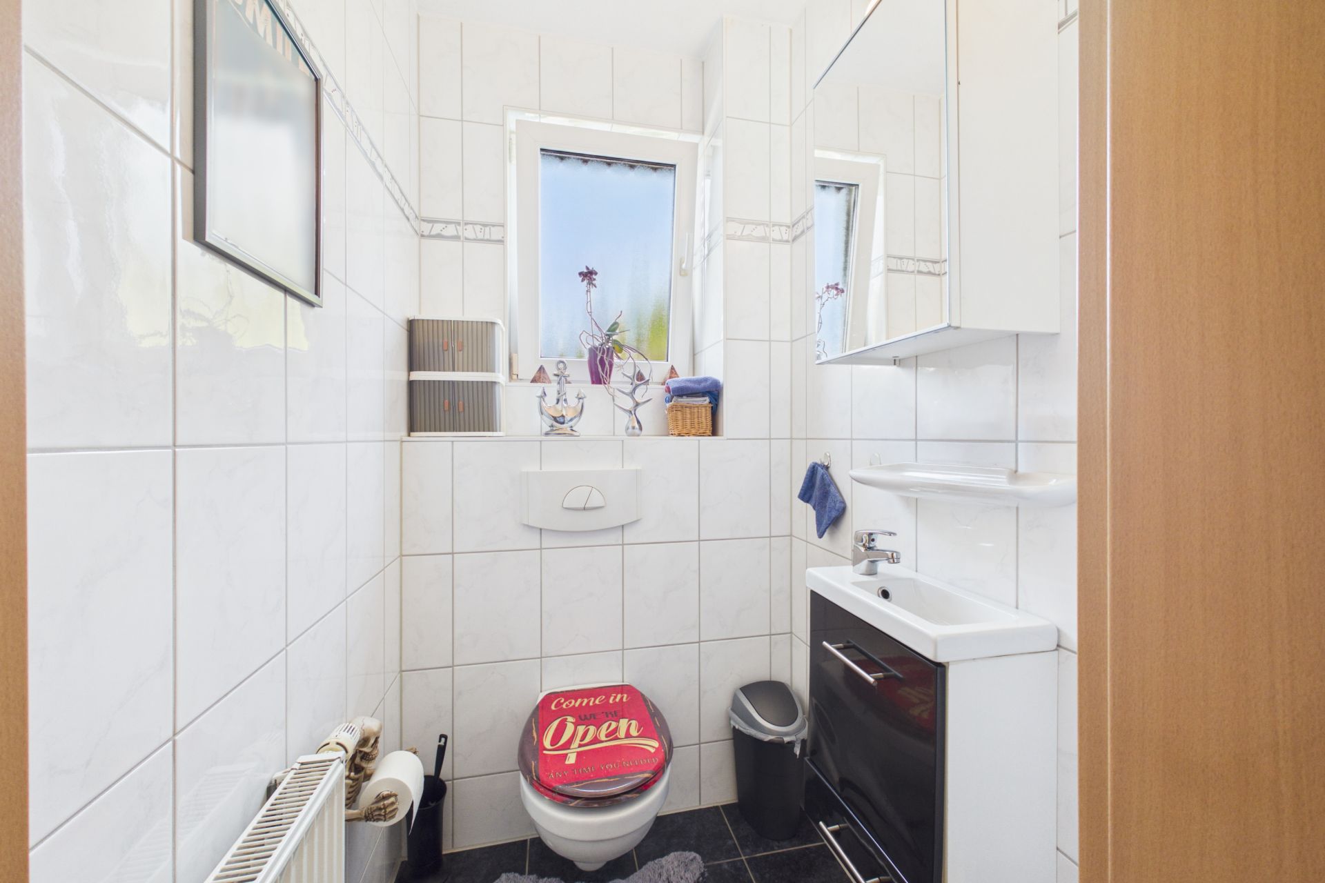 Kleines Bad mit WC, Waschbecken, Spiegel, Fenster und weiß gefliesten Wänden.