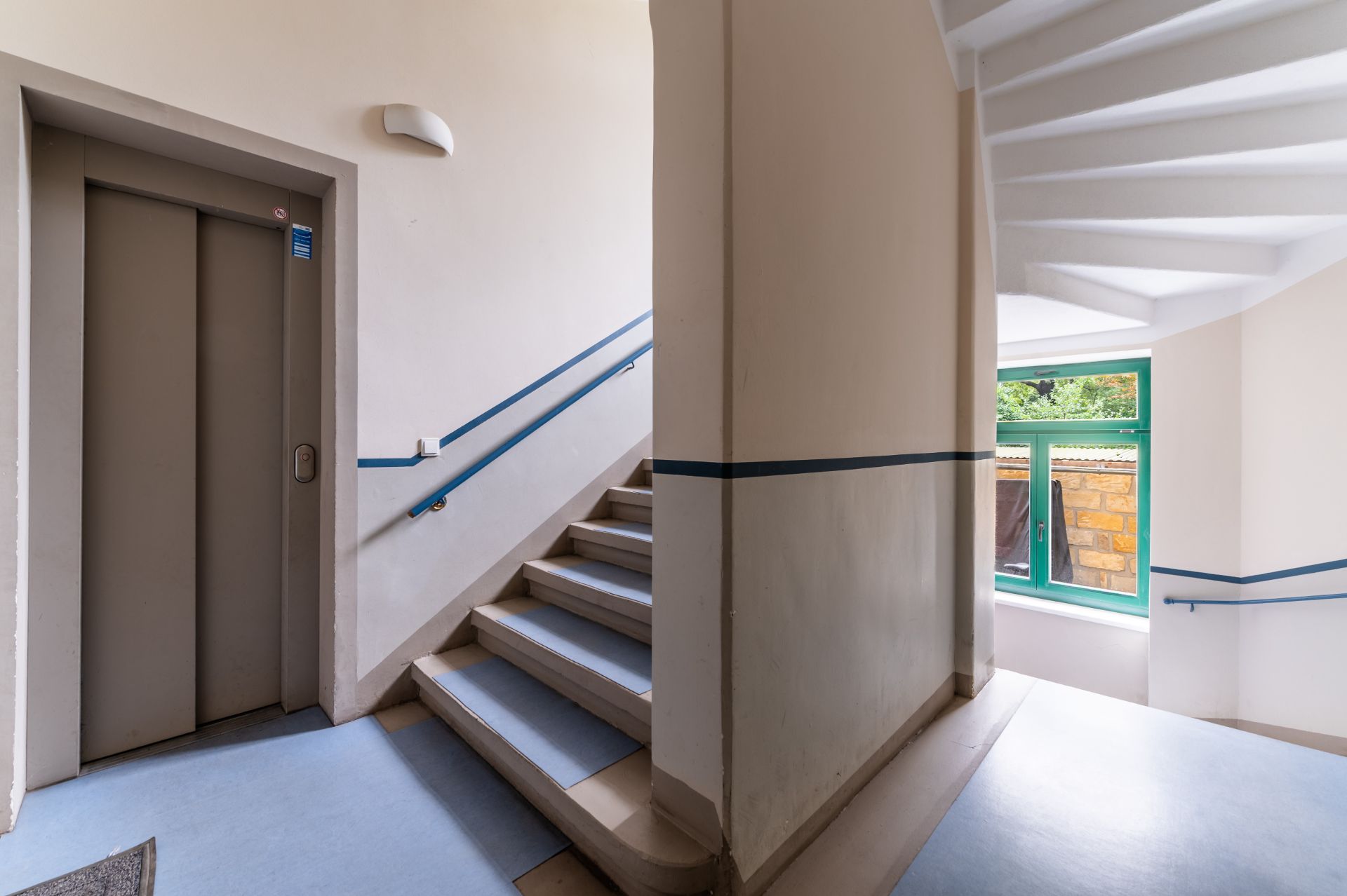Treppenhaus mit Aufzug, blauen Handläufen und Fenster mit Blick nach draußen.