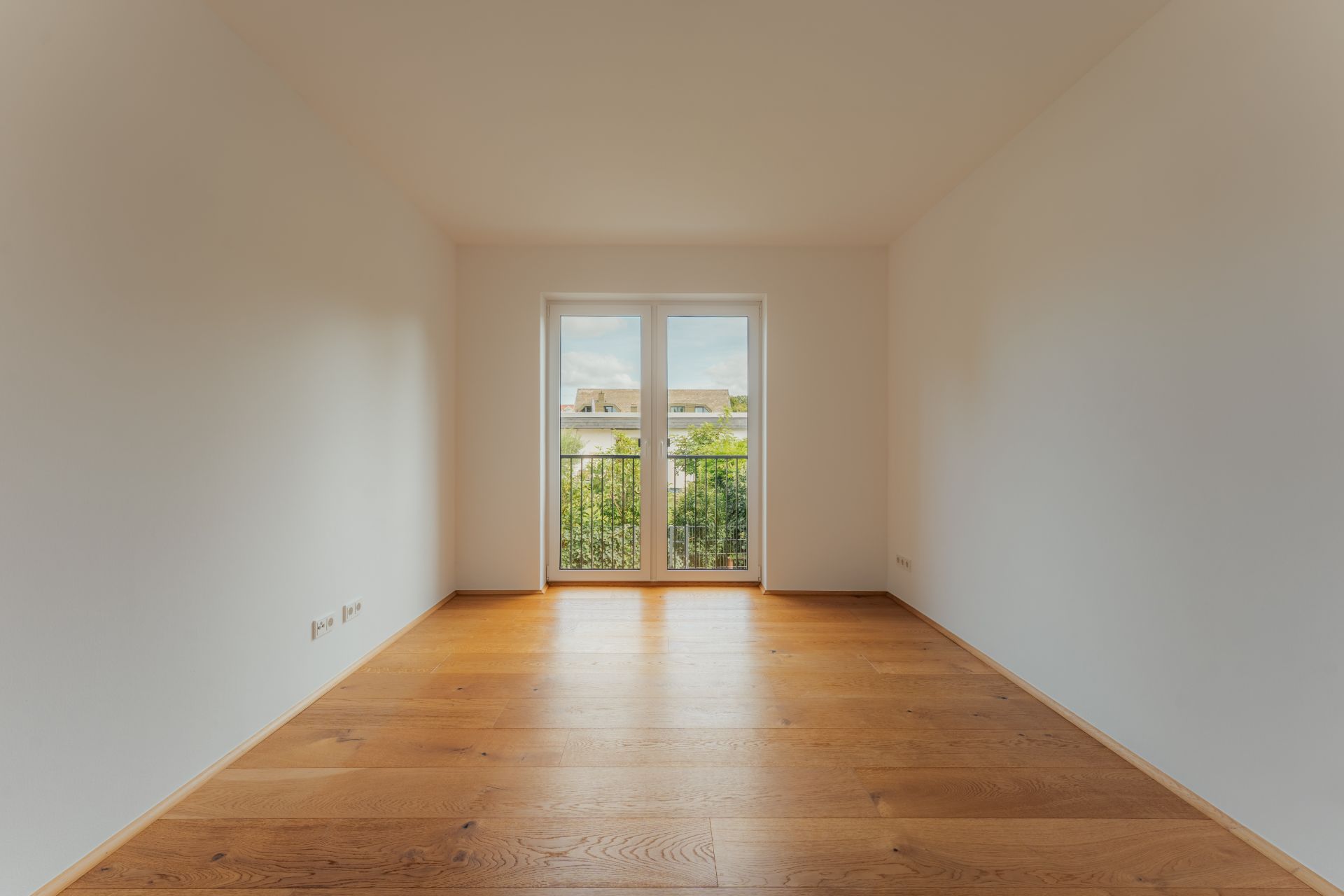 Leerer Raum mit Holzfußboden und Doppeltür zum Balkon, viel Tageslicht.