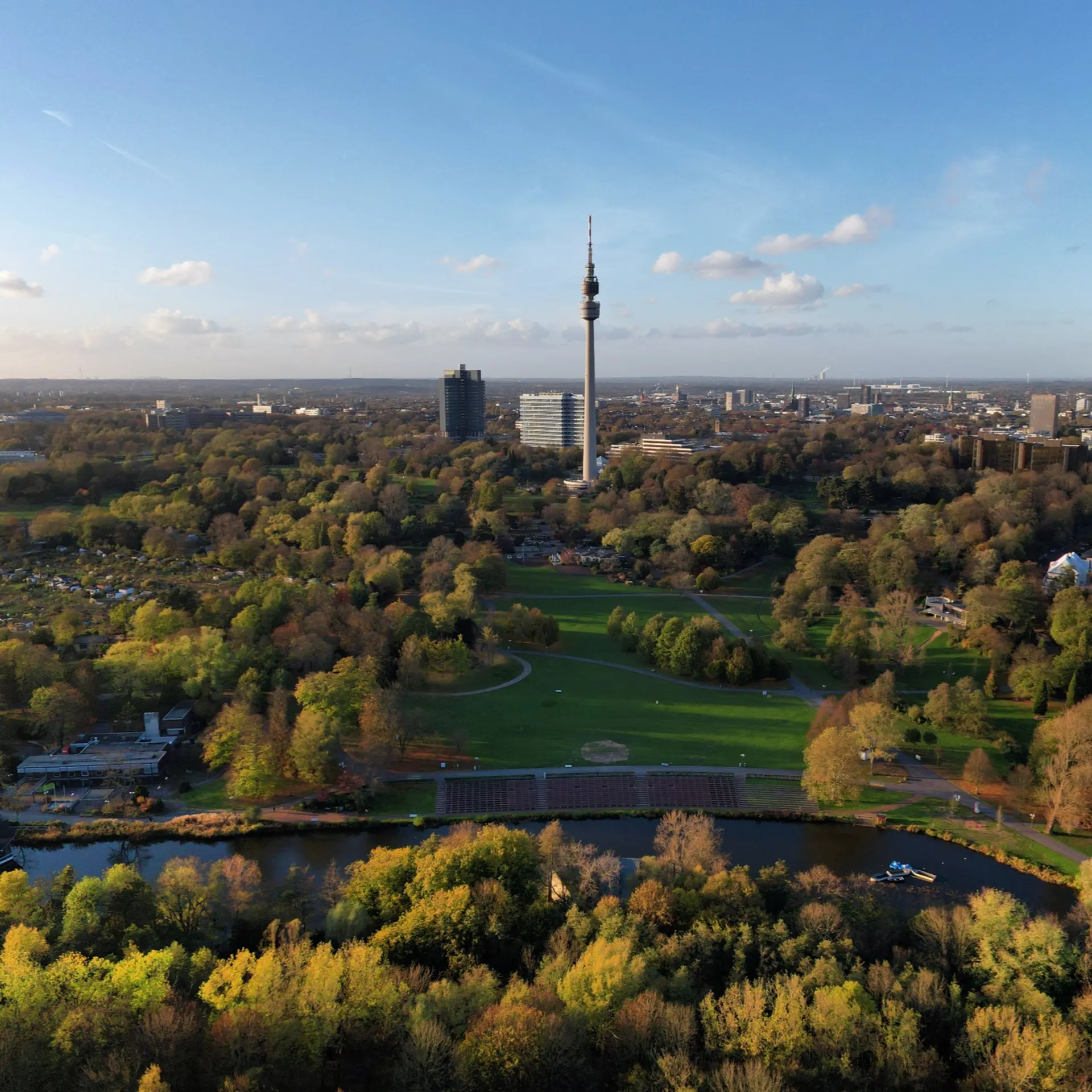 Blick auf einen großen Park mit vielen Bäumen und einem Teich, im Hintergrund ein Fernsehturm.