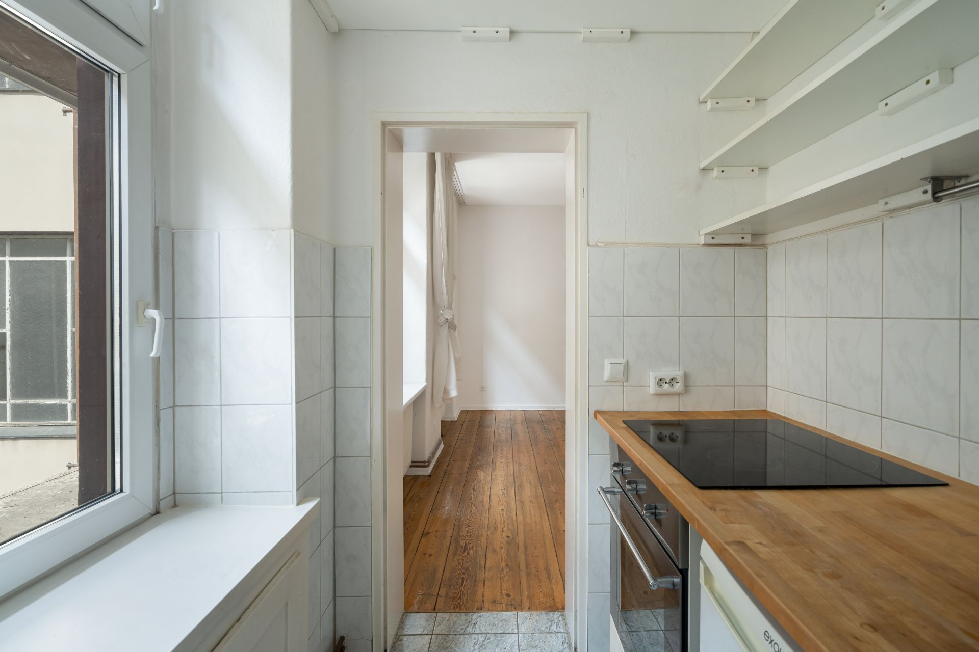 Schmale Küche mit Holzboden, Fenster links, Herd und offenen Regalen, Tür zum hellen Raum.