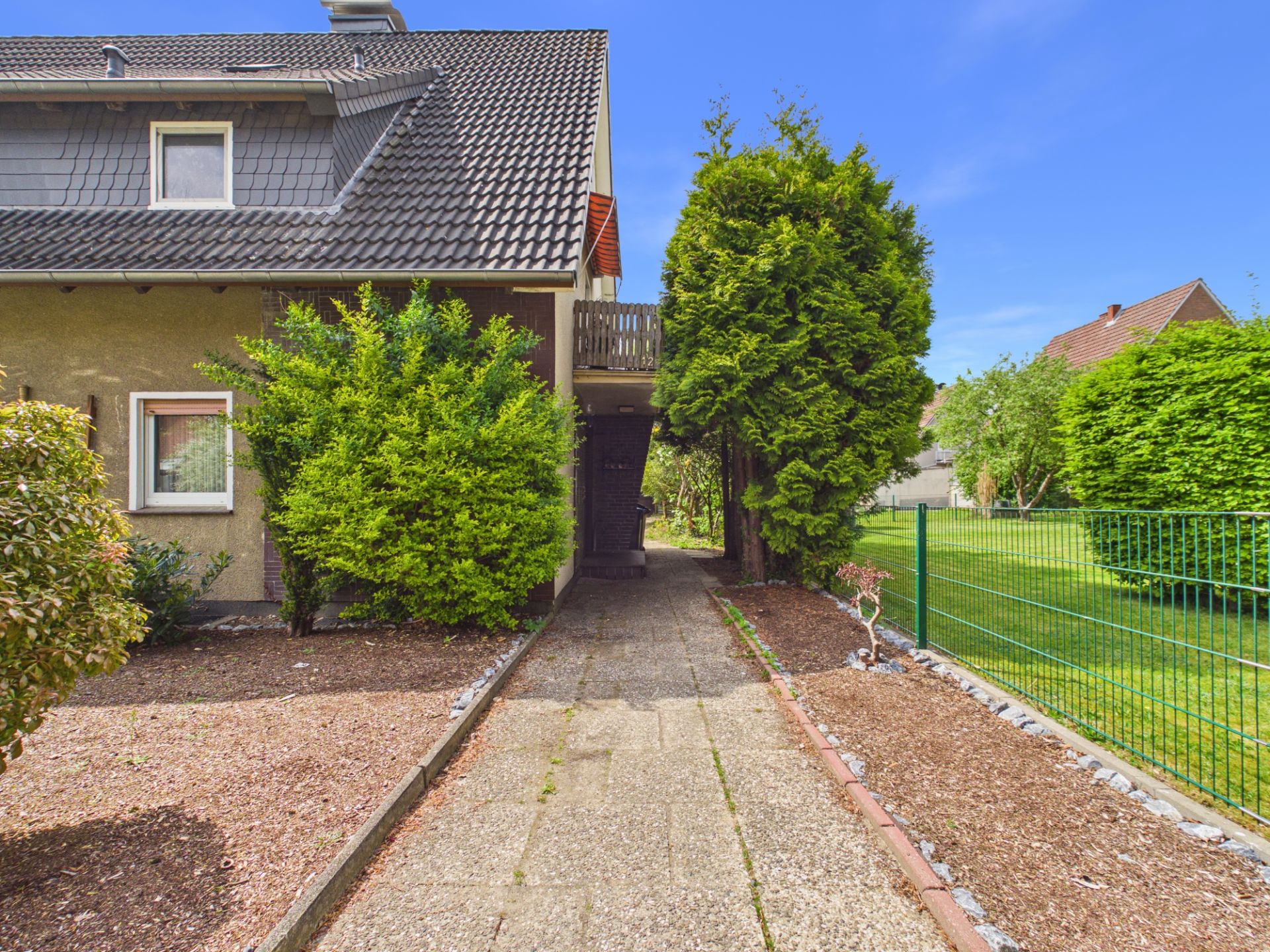 Gepflasterter Weg führt an einem Haus mit Garten und großen Büschen vorbei.