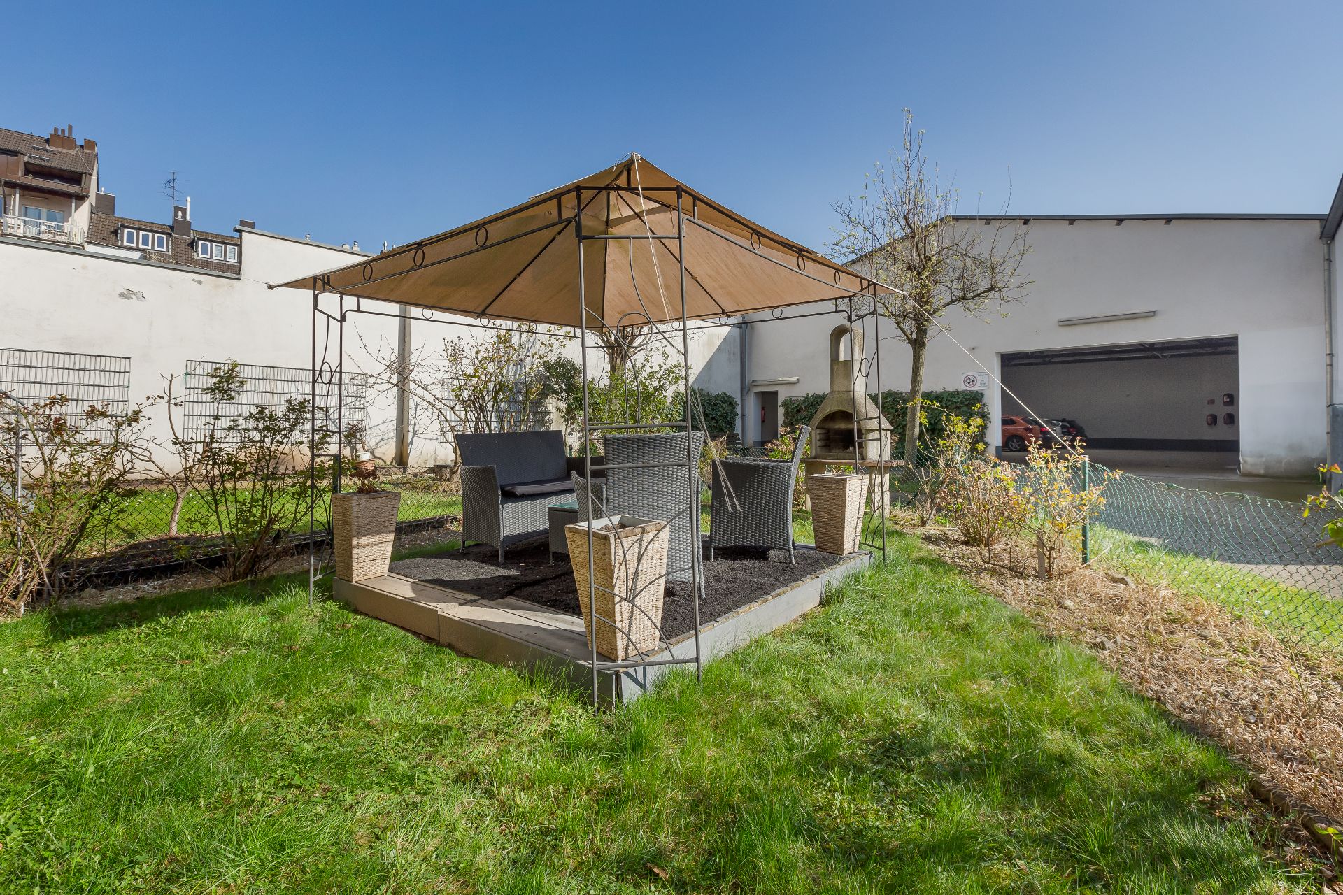 Garten mit Sitzbereich unter Pavillon, Rattanmöbel und Grill im Hintergrund.