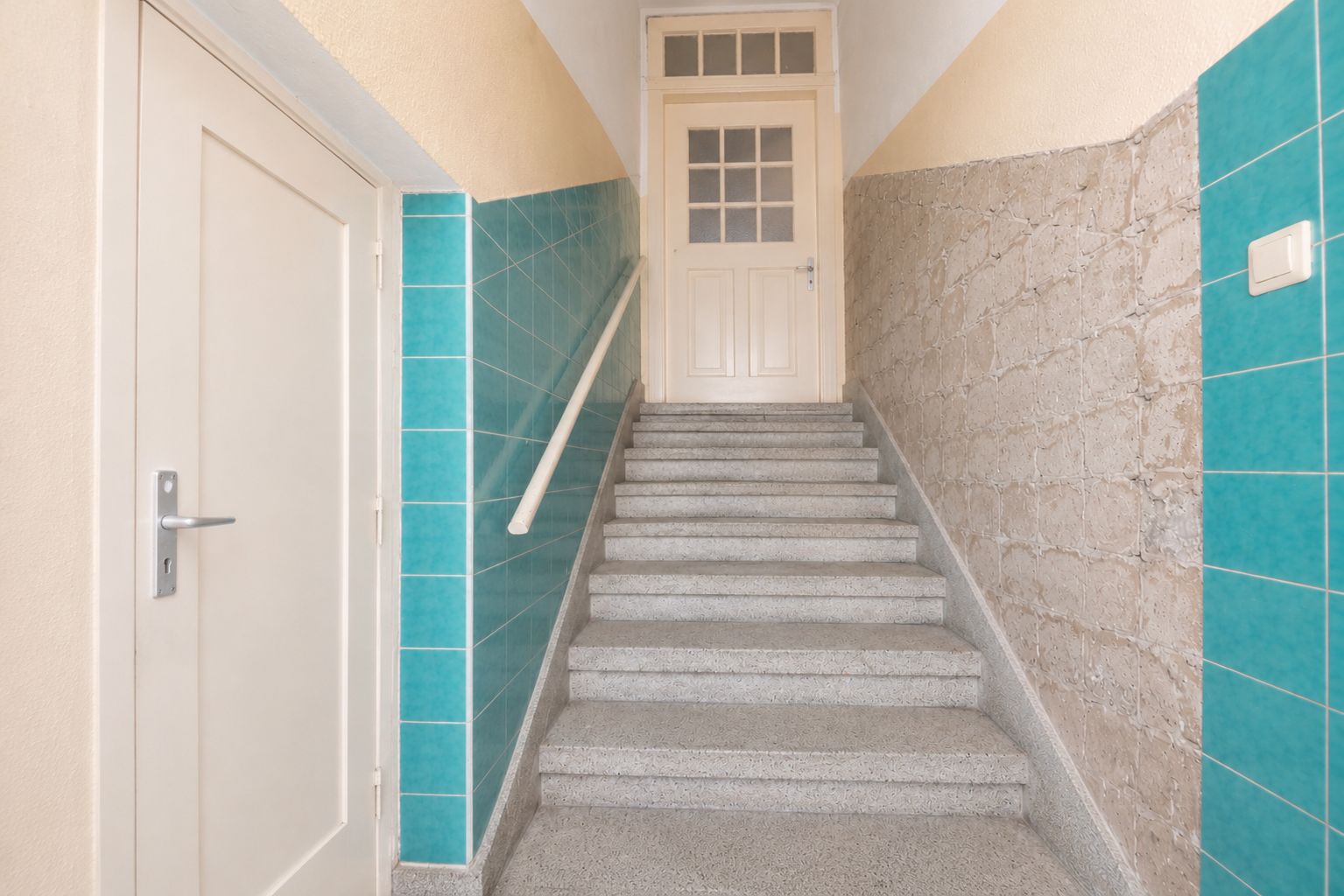 Treppenhaus mit türkisfarbenen Fliesen, Handlauf und einer weißen Tür oben.