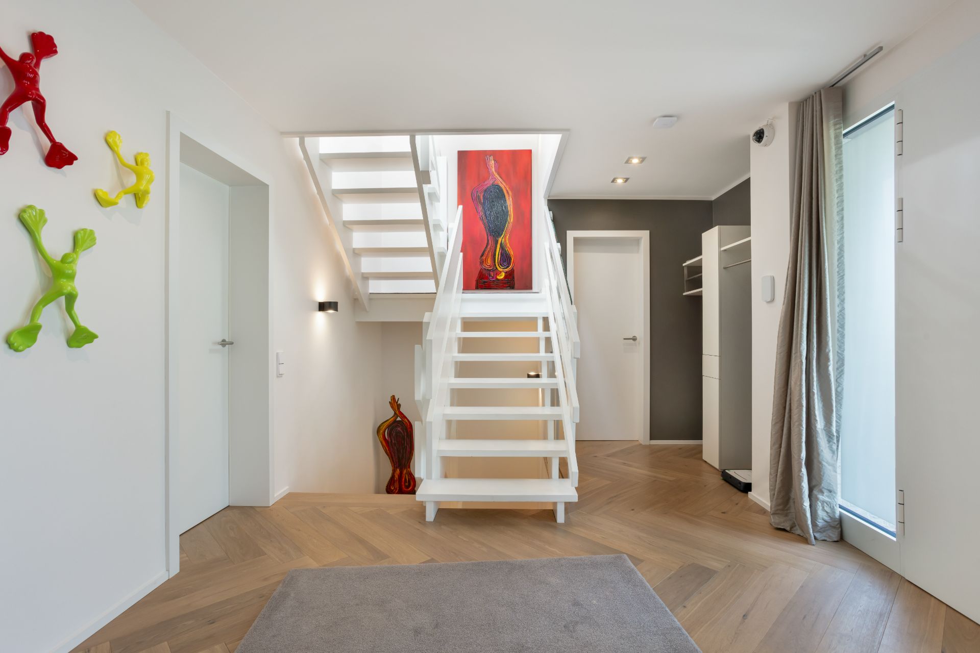 Heller Flur mit weißer Treppe, bunter Wandkunst und Holzboden.