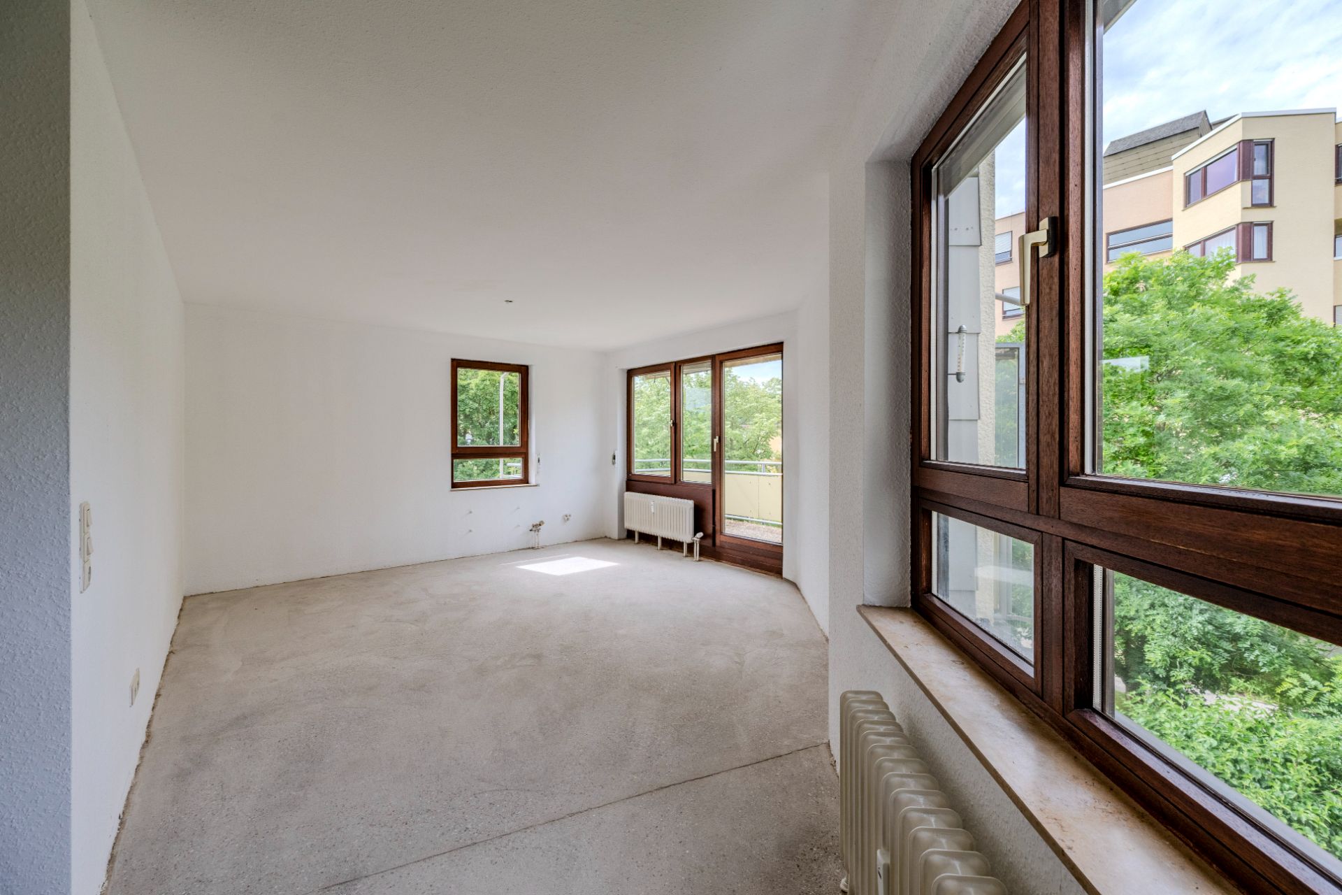 Leeres, helles Zimmer mit großen Fenstern und Blick ins Grüne, Heizkörper unter dem Fenster.