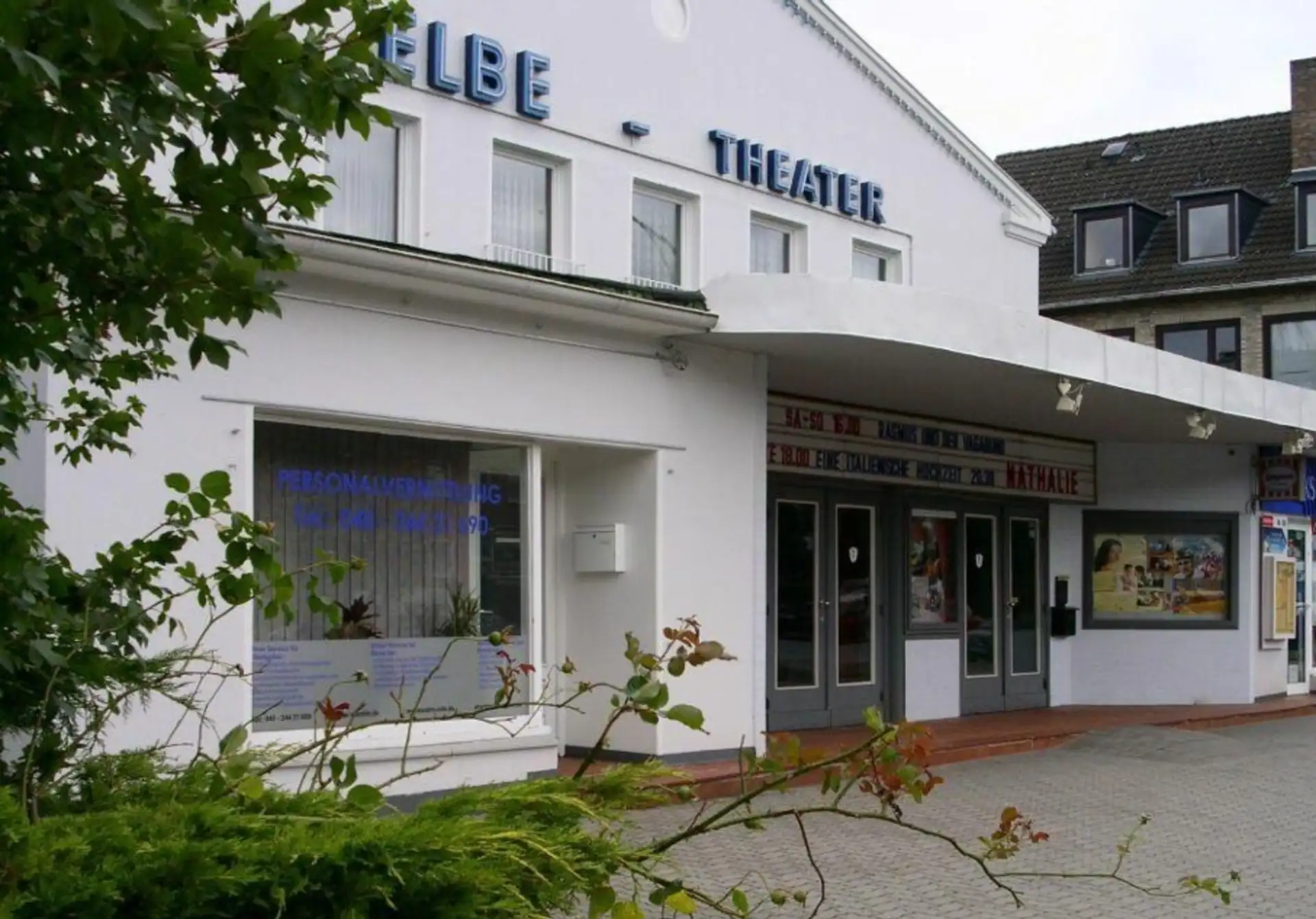 Vorderansicht eines weißen Gebäudes mit Schaufenster, Glastüren und blauem 'THEATER'-Schriftzug.