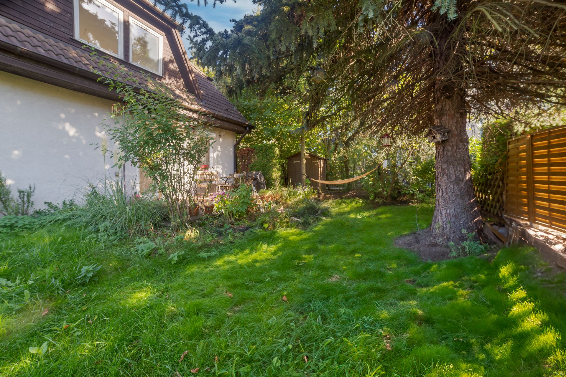 Grüner Garten mit Terrasse, Baum, Hängematte und Gartenhaus neben einem Wohnhaus.
