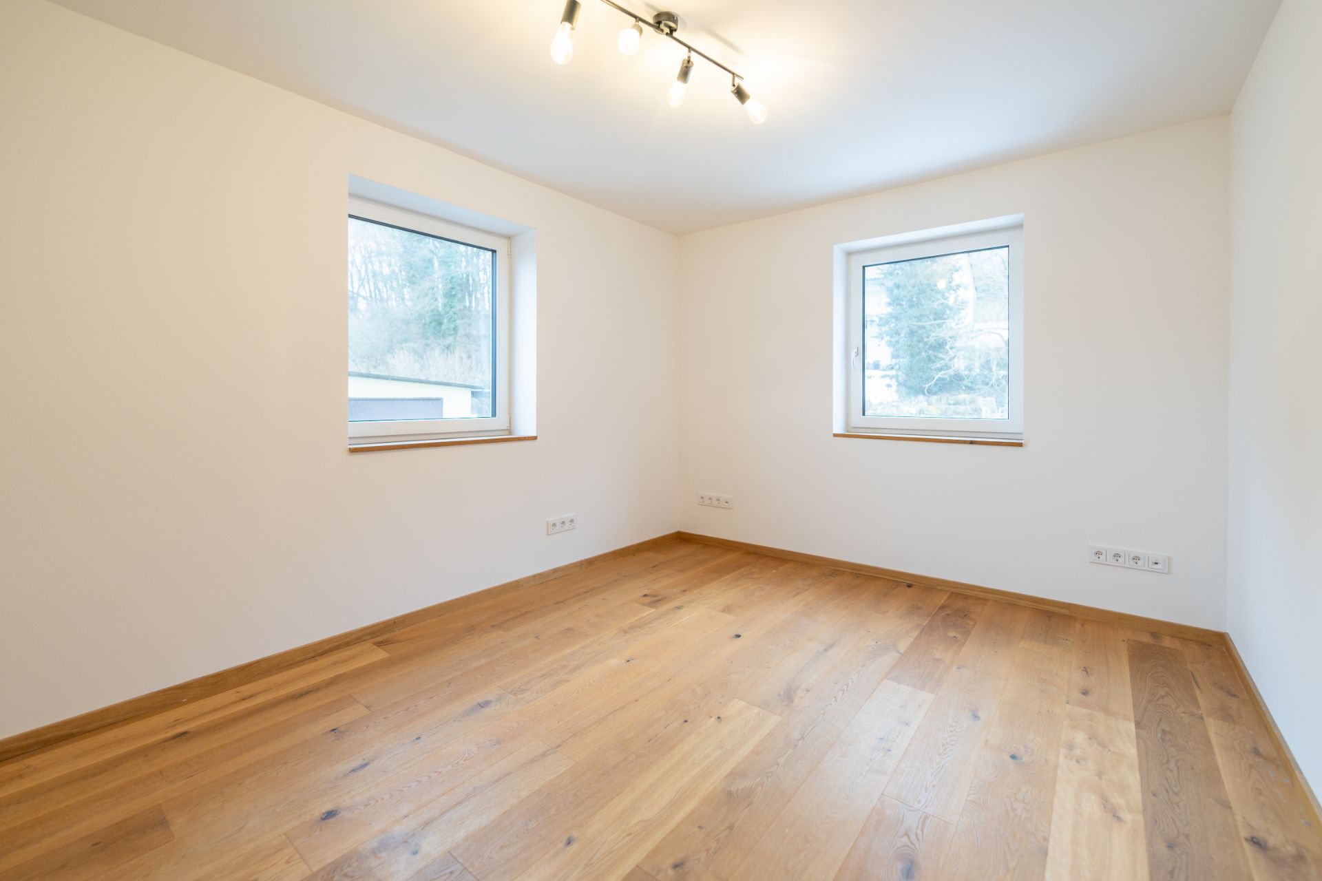 Leeres Zimmer mit Holzfußboden, zwei Fenstern und Deckenleuchte.