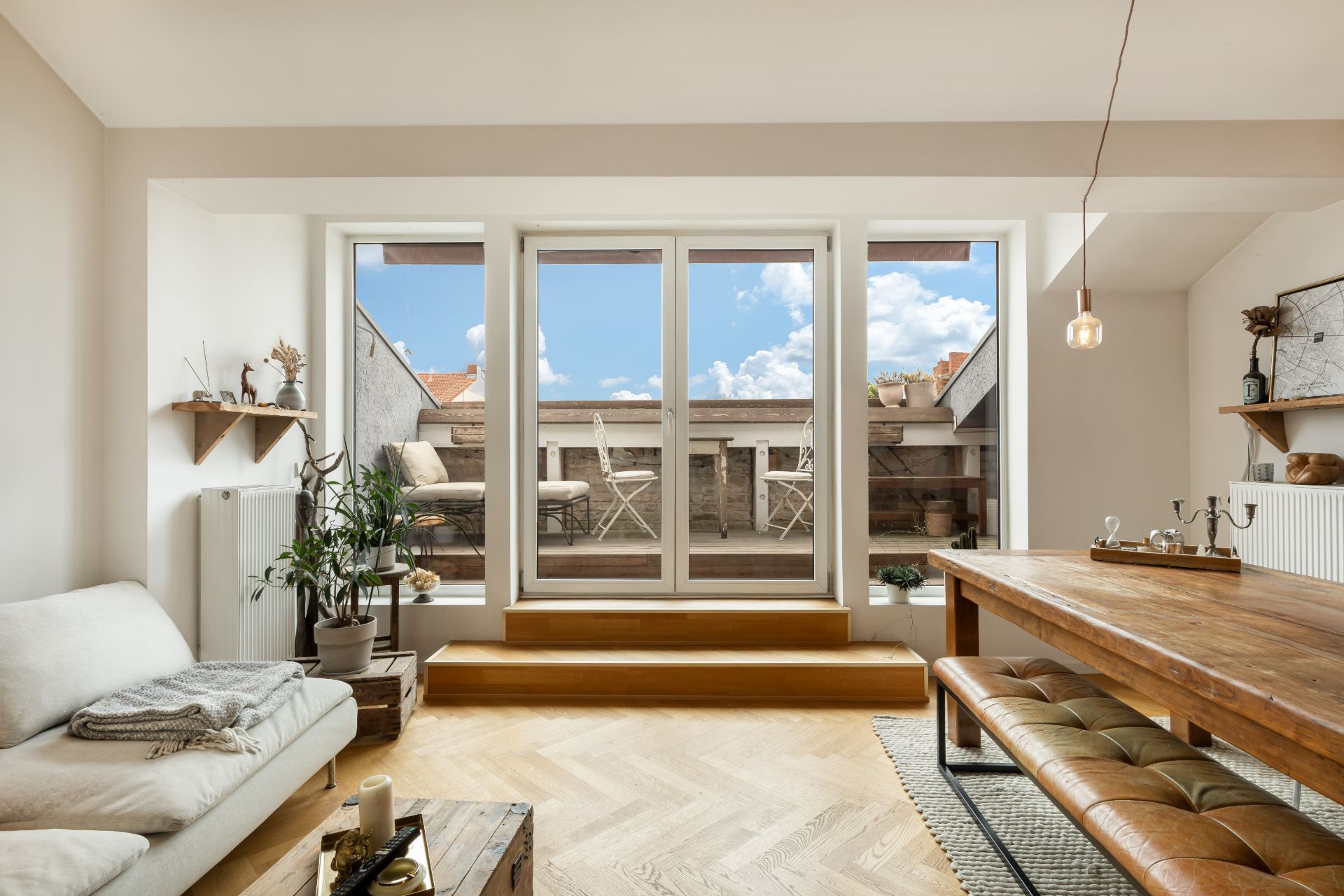 Helles Wohnzimmer mit Sofa, Holztisch, Pflanzen und Blick auf Balkon mit Sitzgelegenheiten.