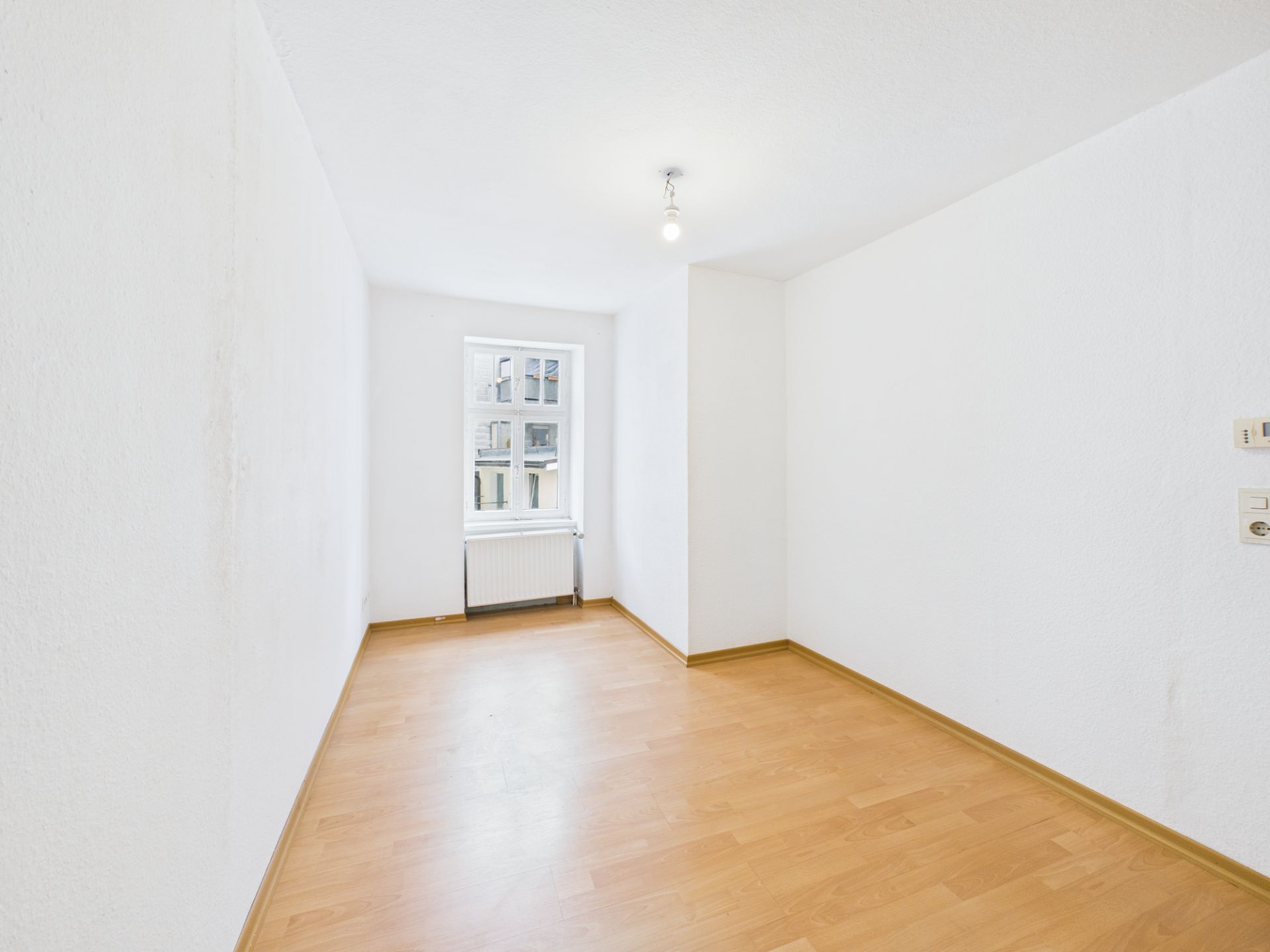 Heller, leerer Raum mit Holzboden, weiß gestrichenen Wänden und einem Fenster mit Heizkörper.