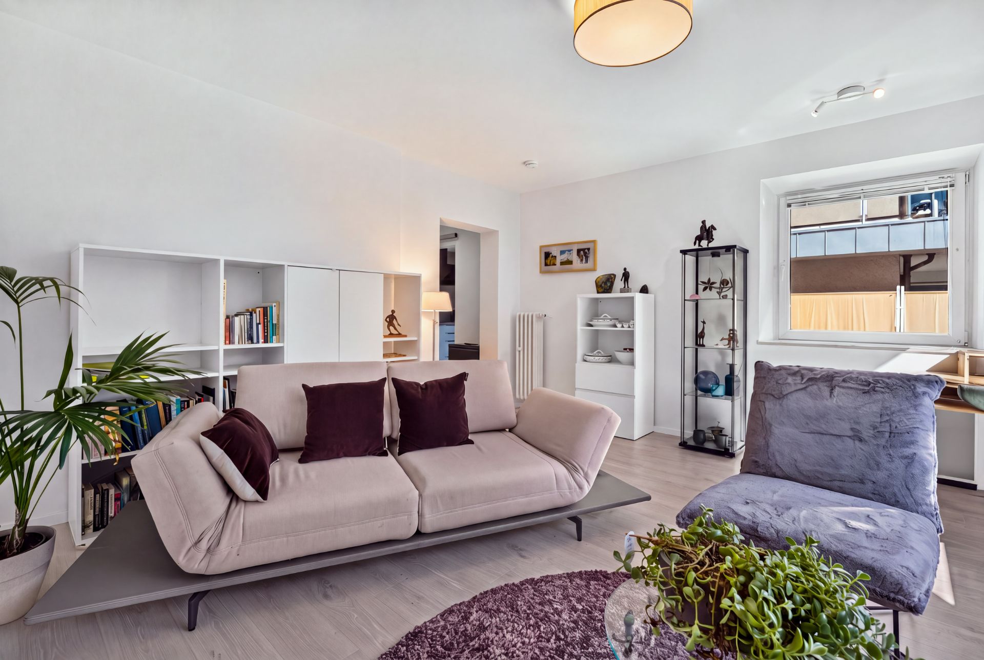 Modernes Wohnzimmer mit beigem Sofa, Sessel, Regal, Pflanzen und großem Fenster.