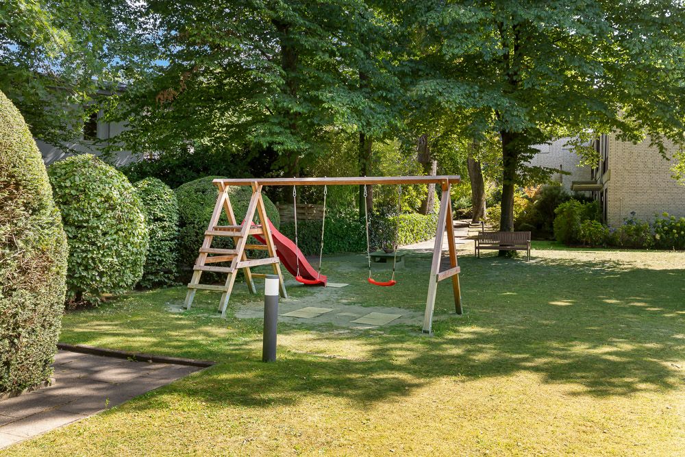 Ein Garten mit Holz-Spielgerüst, zwei Schaukeln und einer roten Rutsche unter Bäumen.