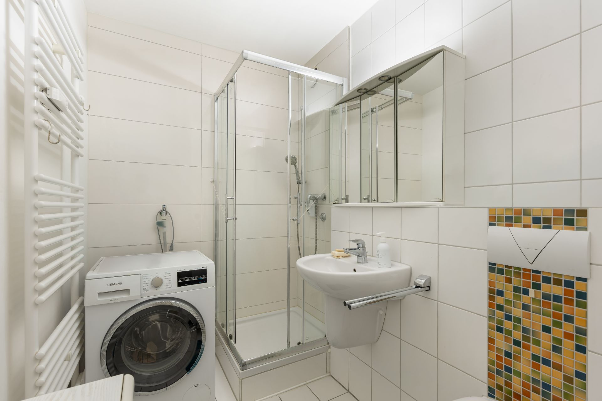 Modernes Badezimmer mit Dusche, Waschmaschine, Waschbecken und farbigem Fliesendekor an der Toilette.