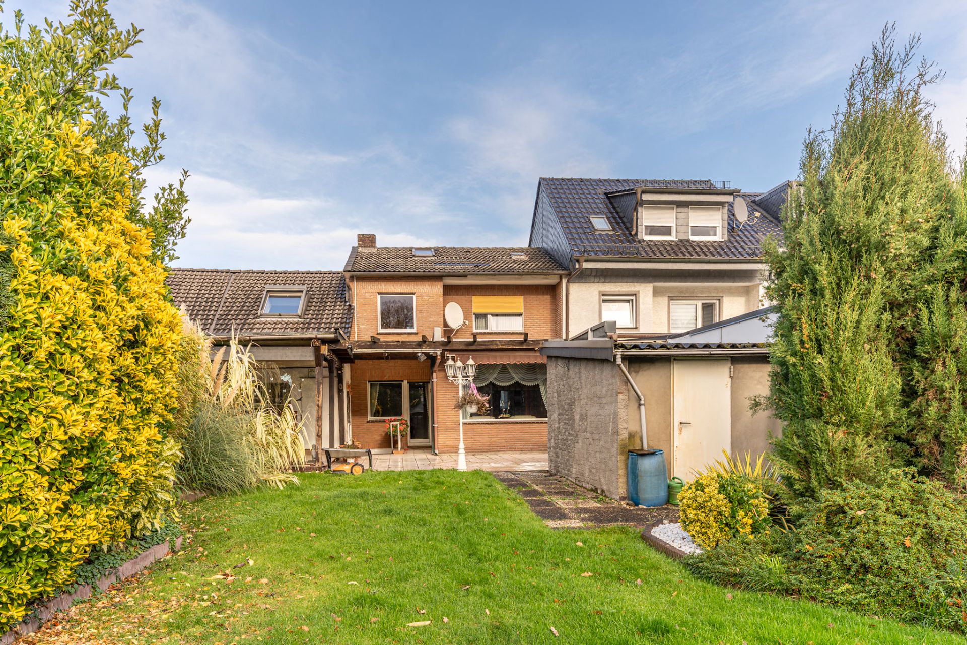 Reihenhaus mit Terrasse, Garten und Schuppen, umgeben von Sträuchern und Bäumen.