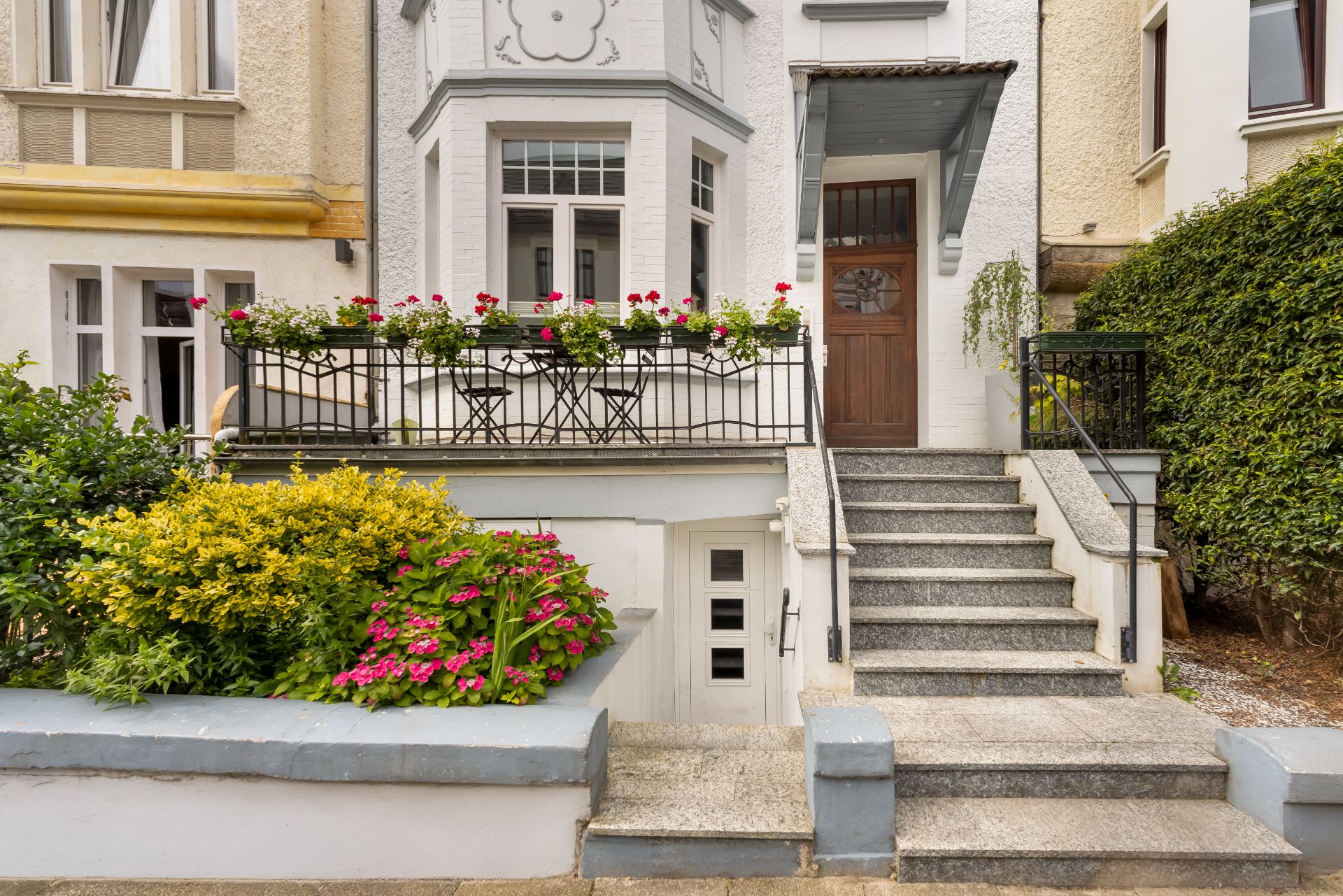 Eingang eines Wohnhauses mit Treppe, Holztür, Blumen und kleiner Terrasse mit Sitzmöbeln.