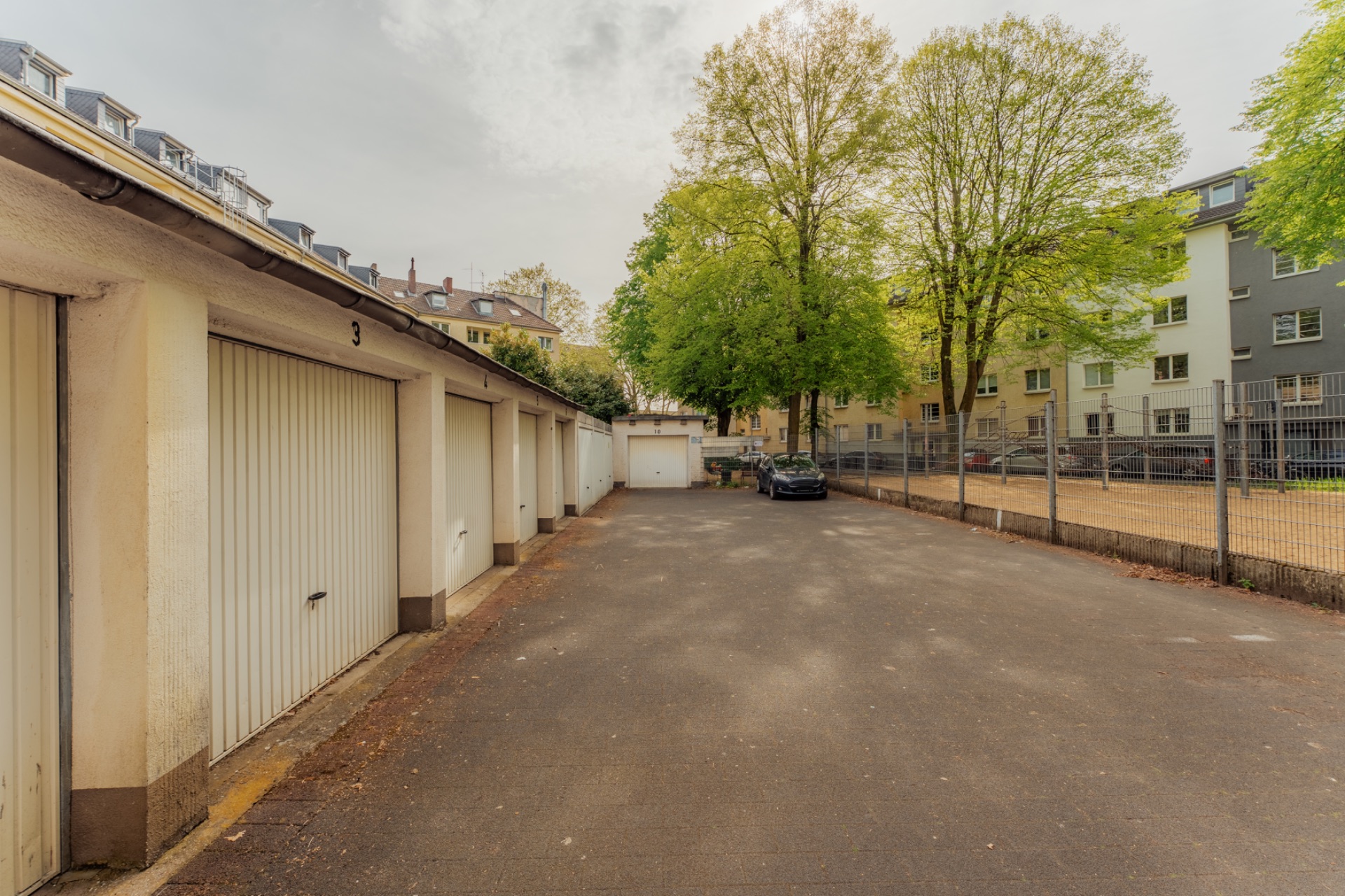 Reihe von Garagen mit geschlossener Tür, daneben ein Parkplatz und Bäume im Hintergrund.