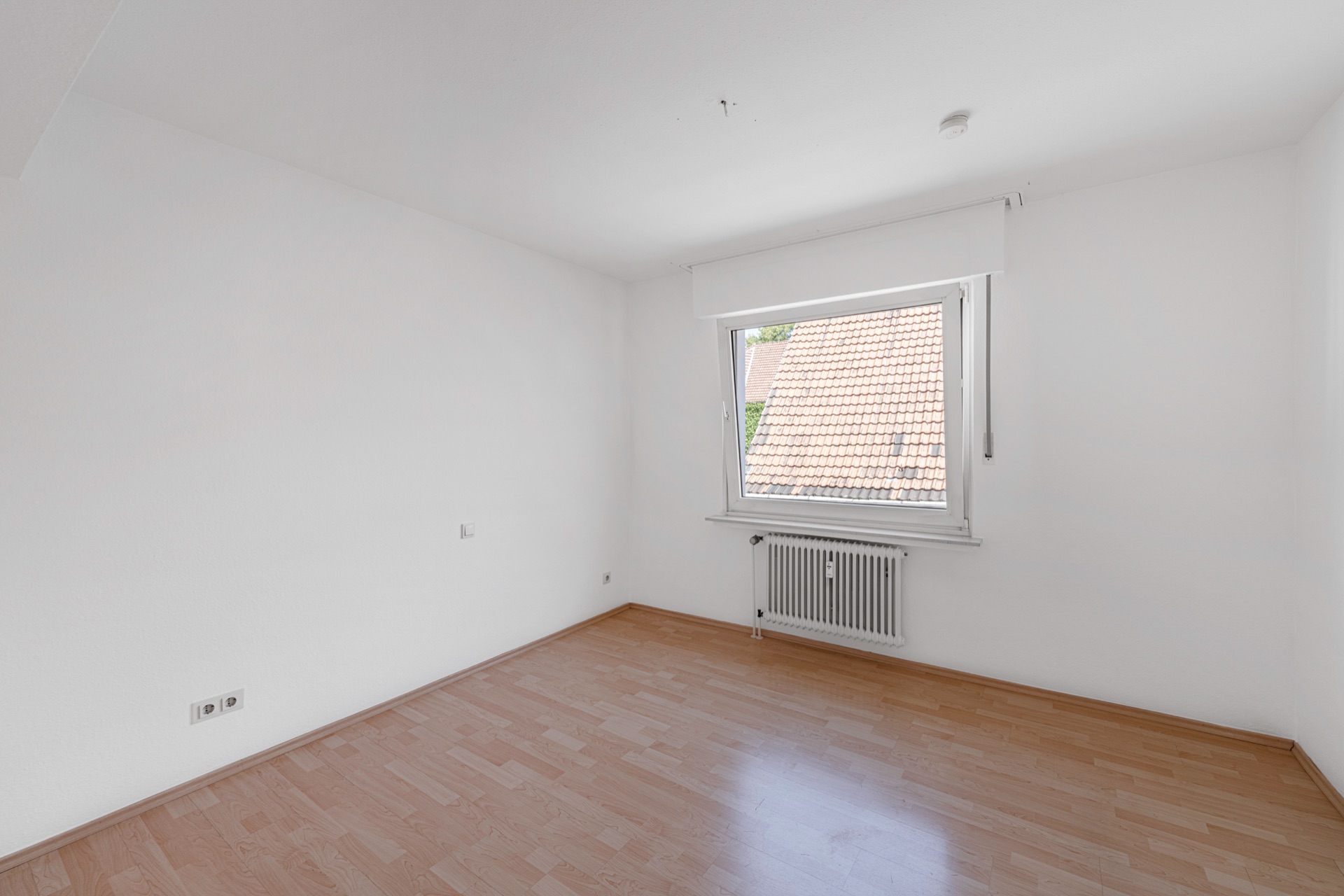 Leerer Raum mit weißem Wänden, Laminatboden und Fenster mit Blick auf ein Ziegeldach.