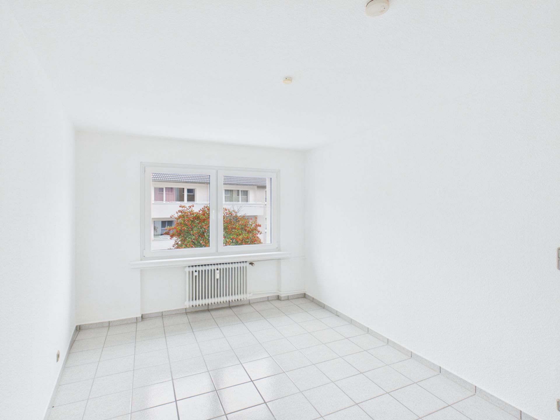 Helles, leeres Zimmer mit weißen Wänden, Fliesenboden und einem Fenster mit Blick nach draußen.