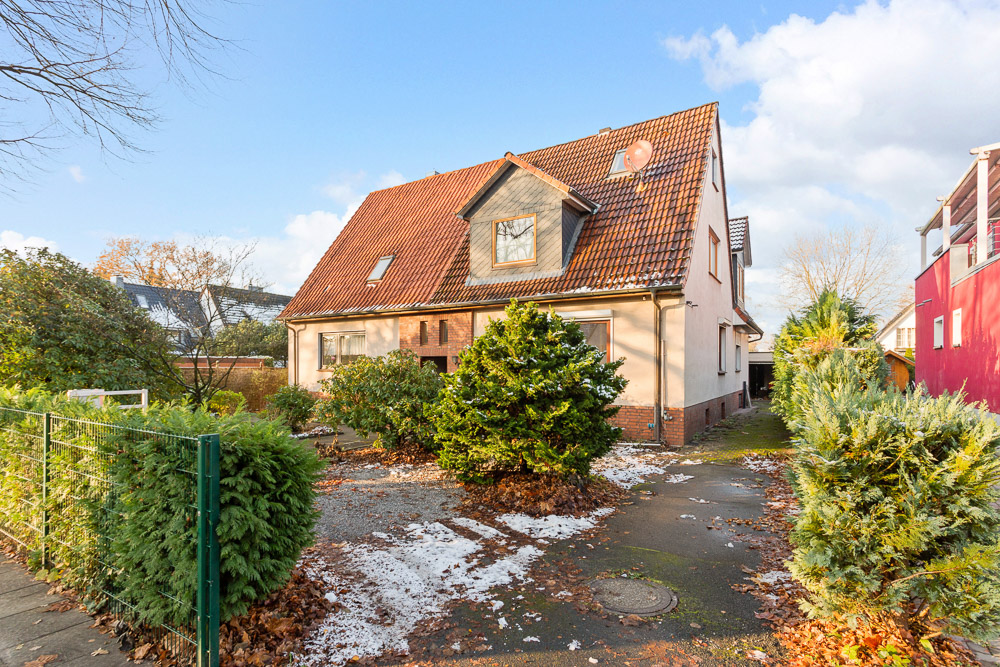 Ein Wohnhaus mit rotem Satteldach, Vorgarten und einigen Büschen, teils mit Schnee bedeckt.