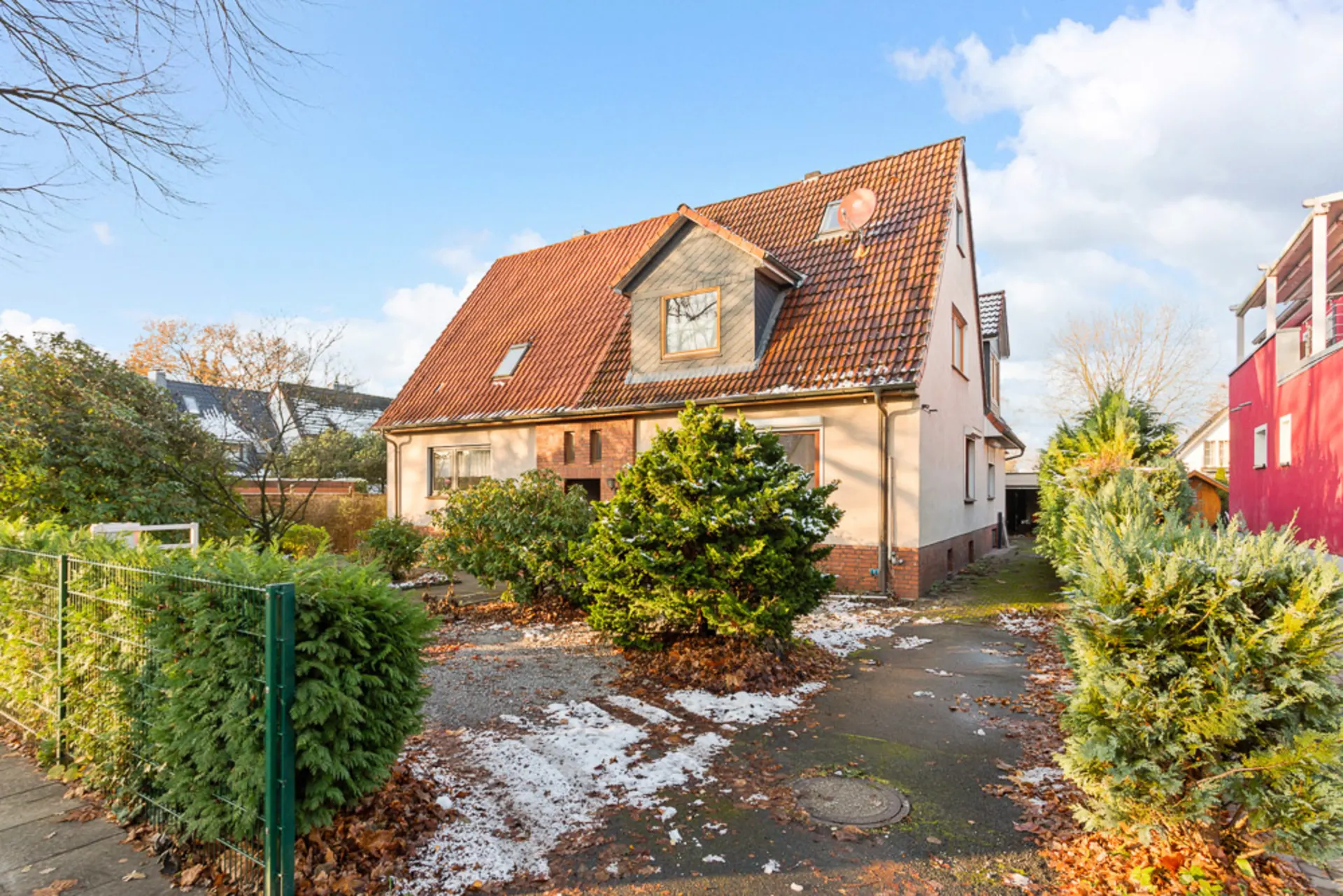 Ein Wohnhaus mit rotem Satteldach, Vorgarten und einigen Büschen, teils mit Schnee bedeckt.