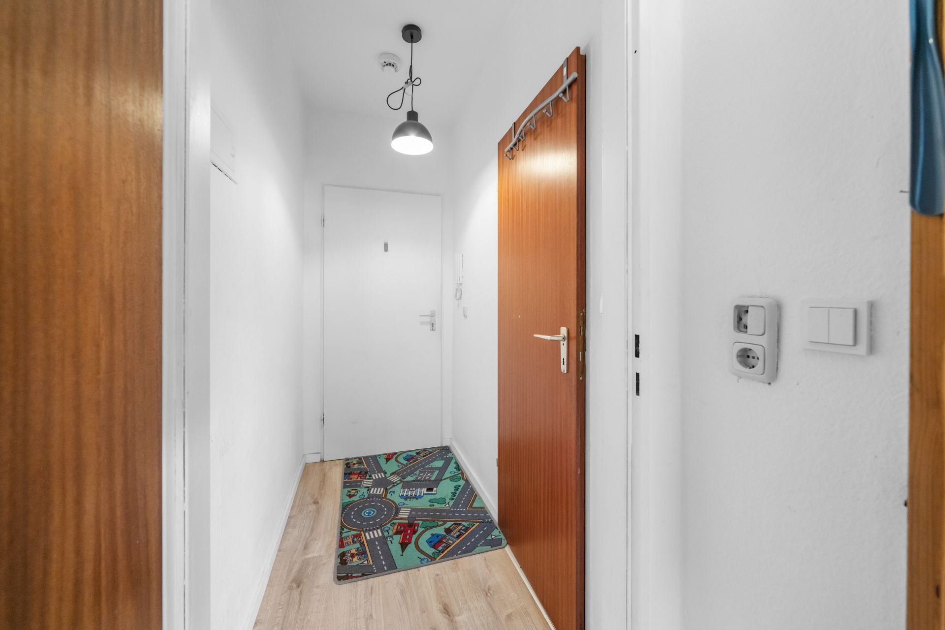 Heller Flur mit Holztüren, weißer Wand, bunter Teppich und Hängelampe.