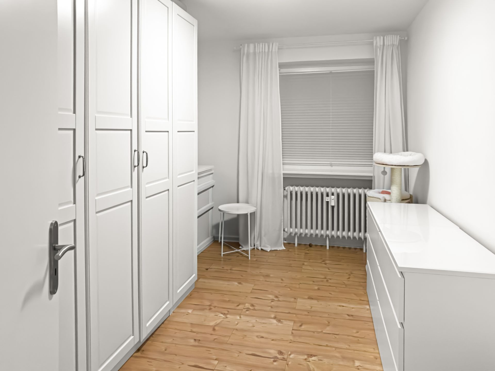 Helles Zimmer mit weißen Schränken, Kommode, Heizkörper und Holzfußboden.