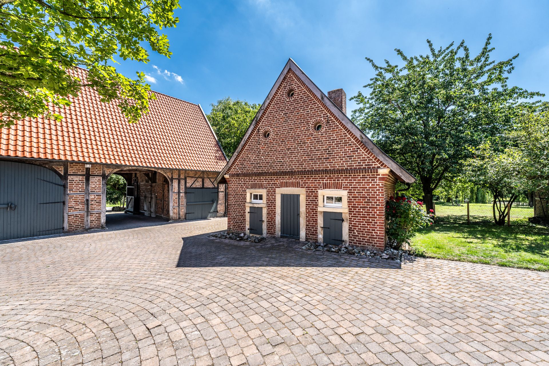 Backstein-Gebäude mit spitzem Dach, gepflastertem Hof und Bäumen im Hintergrund.