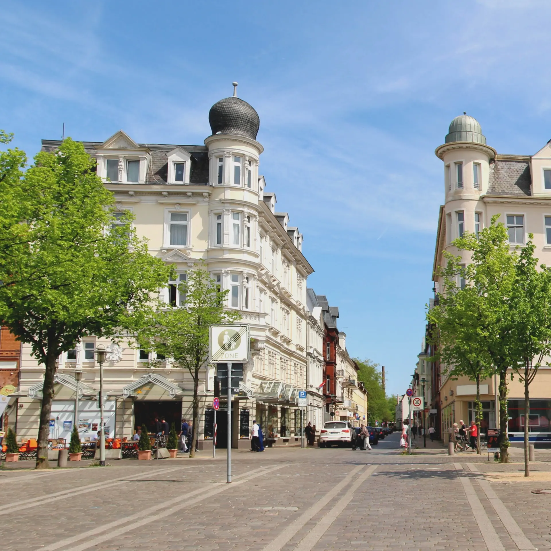 Straße mit historischen Wohngebäuden, Bäumen und Gehweg, mehrere Menschen und Autos sichtbar.