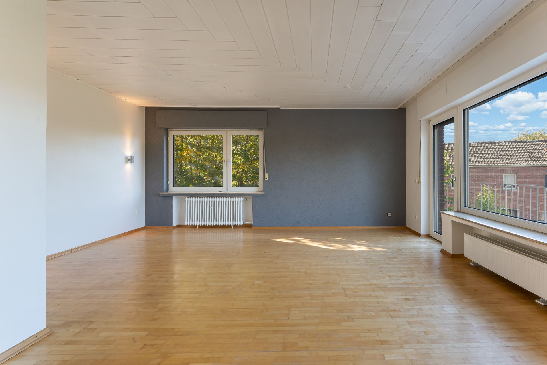 Großes, leeres Wohnzimmer mit Parkettboden, weißen Wänden und großen Fenstern.